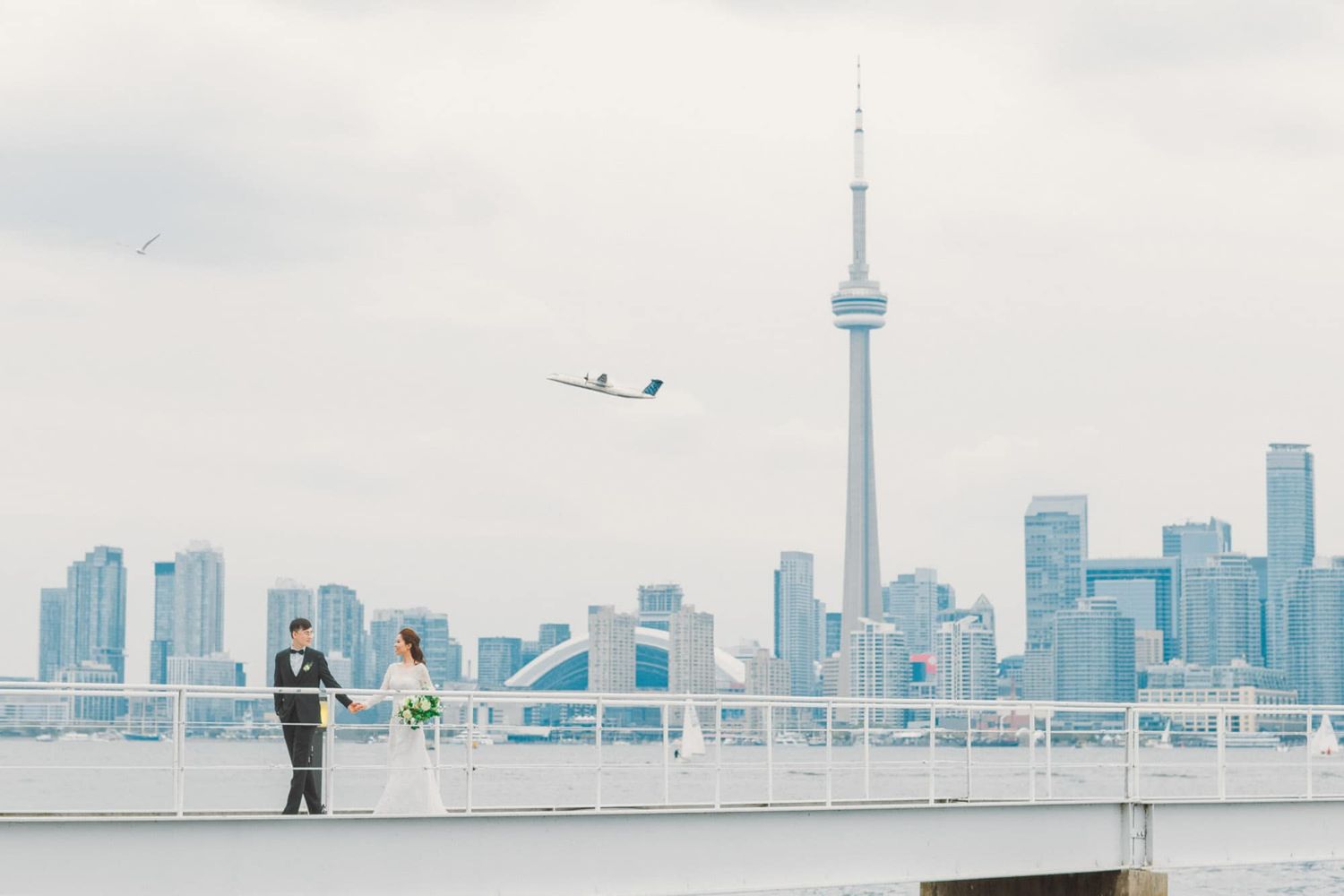 Bride and groom hold hands on a waterfront pier with the Toronto skyline and CN Tower in the background.