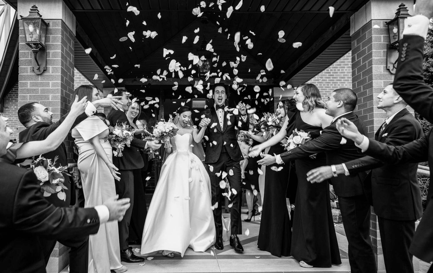 Black and white photo of newlyweds walking through flower petal toss outside wedding venue entrance.