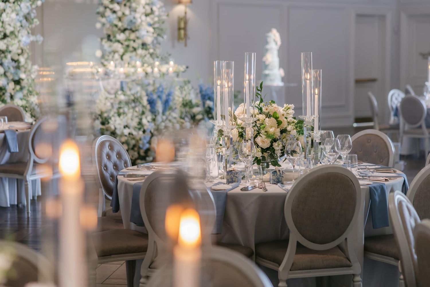 Elegant wedding reception with round tables, blue linens, tall candles, and white floral centerpieces in a grand ballroom.