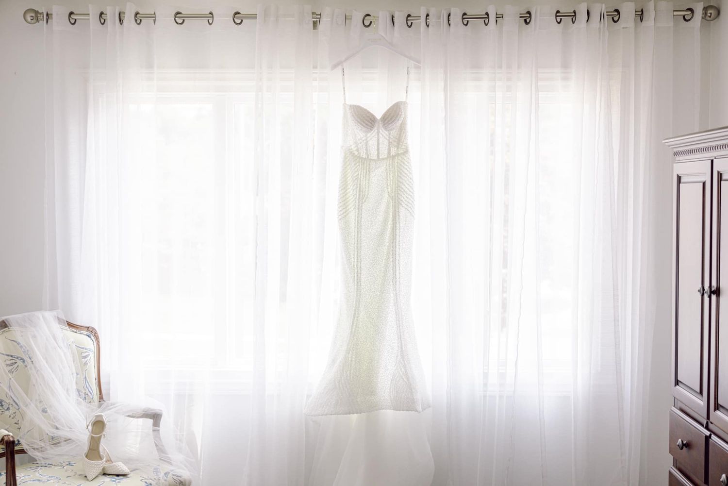 Elegant strapless white mermaid wedding dress hanging against sheer white curtains in bright bridal suite.