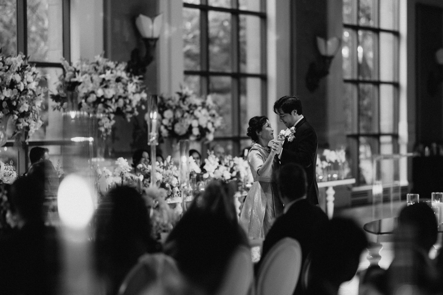 Bride and groom share a romantic first dance in an elegant ballroom with floral centerpieces as wedding guests watch in black and white.