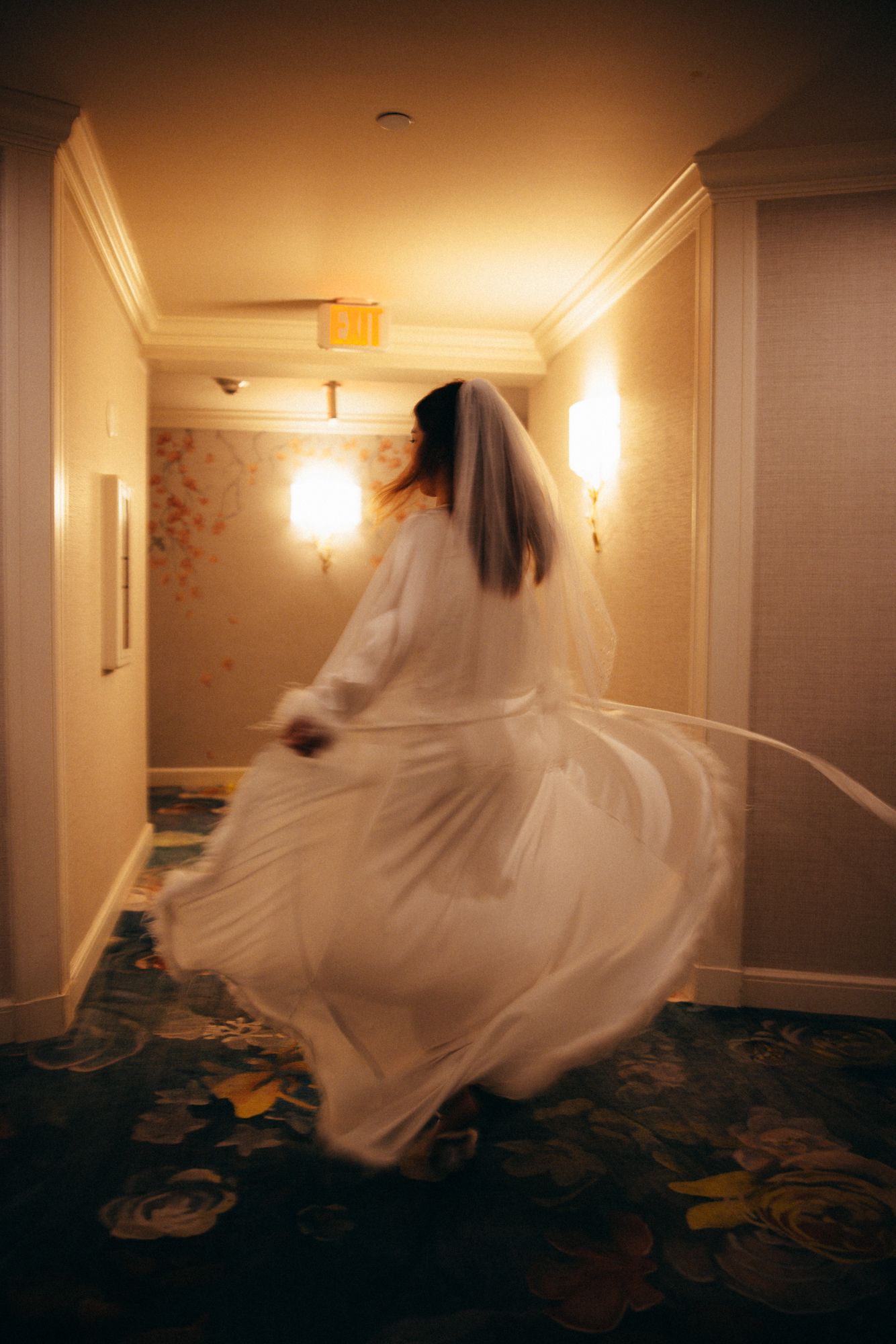 Bride in white wedding dress and veil twirling in hotel corridor with warm golden wall sconces.