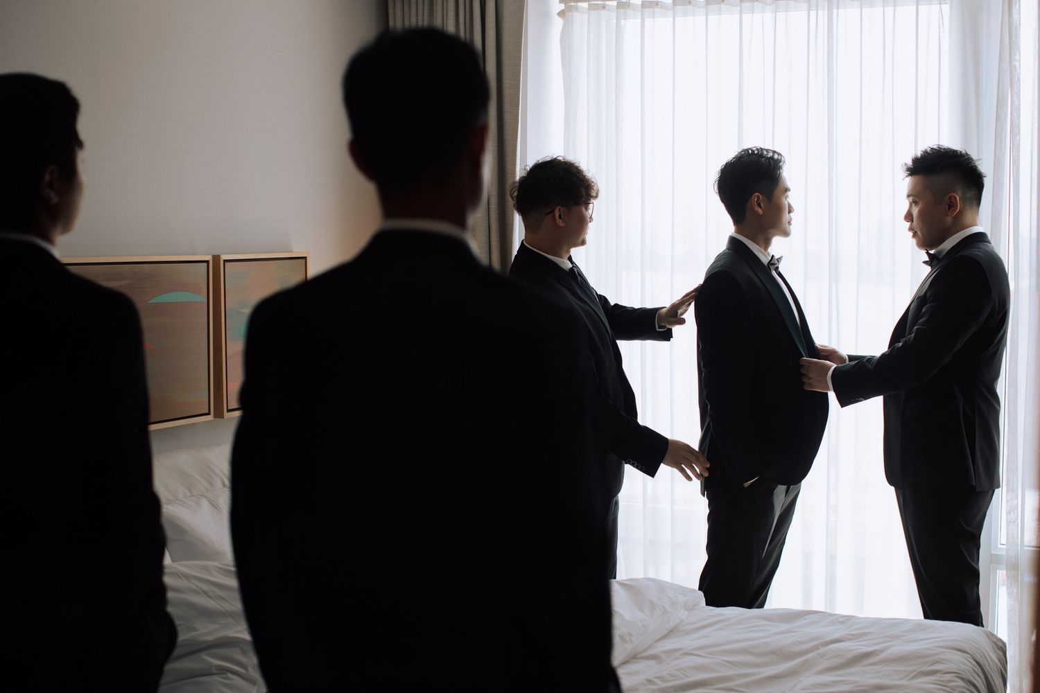 Group of groomsmen in black suits assisting groom while getting ready in hotel room.