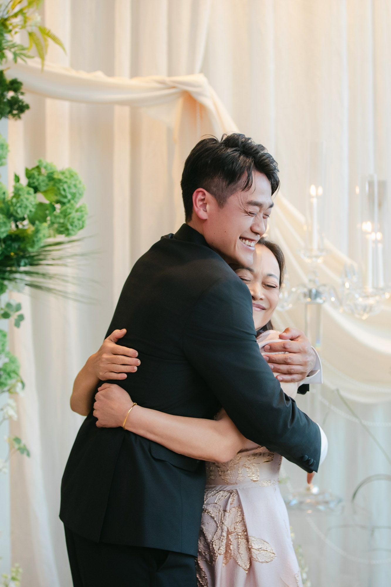 Groom in black tuxedo sharing a warm embrace with a woman in a floral dress at the wedding.