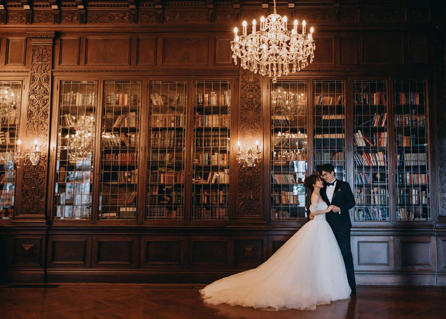 Couple embraces romantically in a grand wood-paneled library beneath a glowing chandelier.