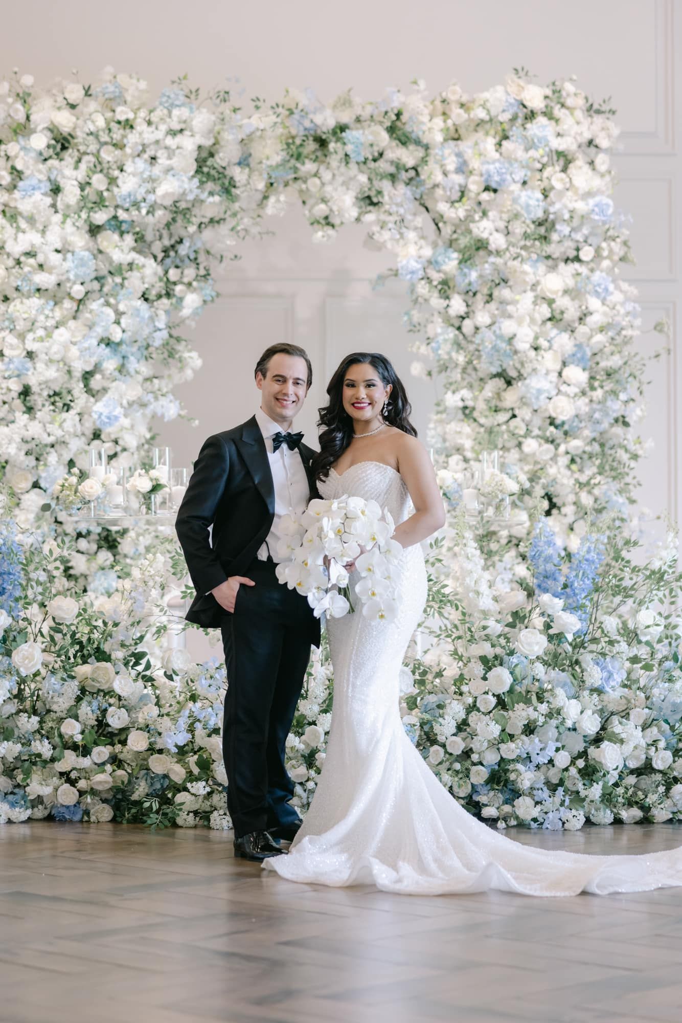 Bride in mermaid gown and groom in tuxedo stand before a stunning blue and white floral arch backdrop.