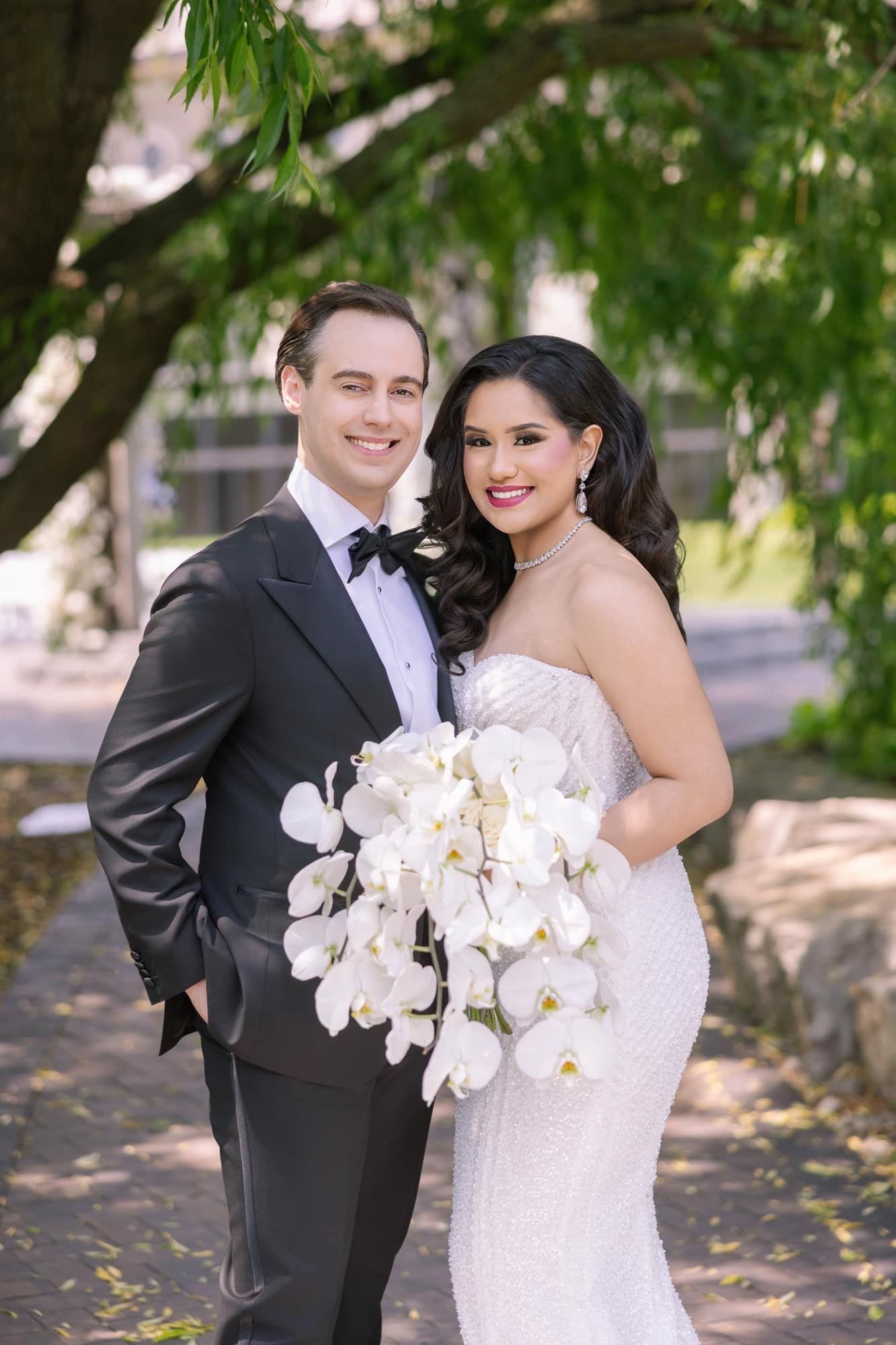 Smiling newlywed couple posing outdoors under green trees, bride holding cascading white orchid bouquet.
