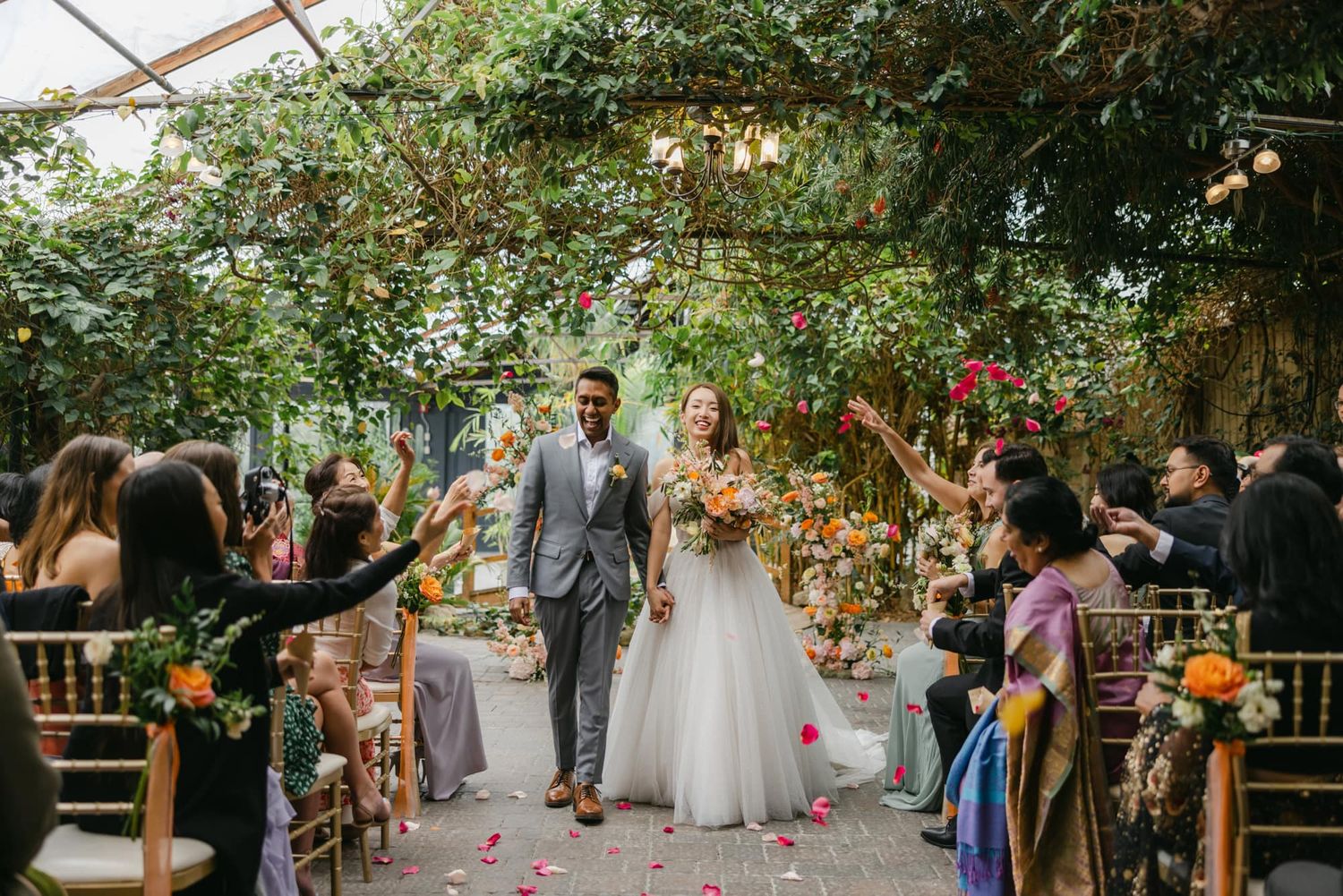 Joyful couple exits outdoor garden wedding ceremony as guests toss colorful flower petals.