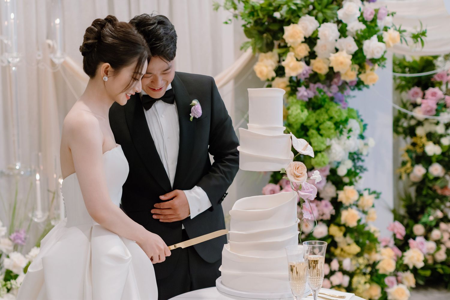 Bride and groom cutting a tall white tiered wedding cake surrounded by lush floral arrangements.