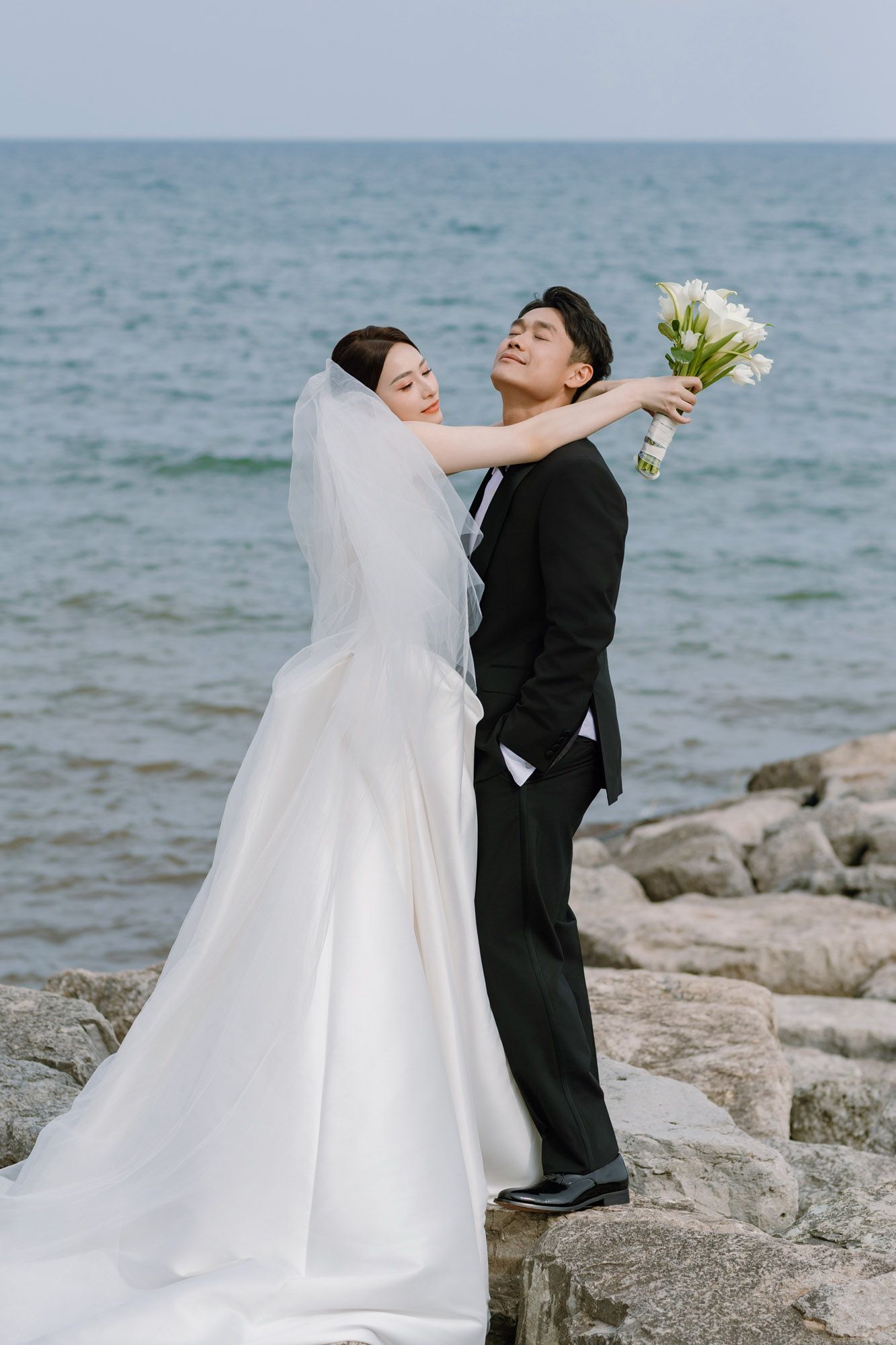 Bride in flowing white gown and veil embraces groom on rocky oceanside wedding photo.