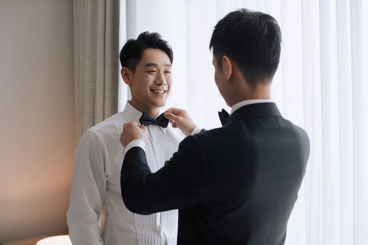 Groomsman adjusting bow tie on smiling Asian groom in white tuxedo shirt before wedding ceremony.