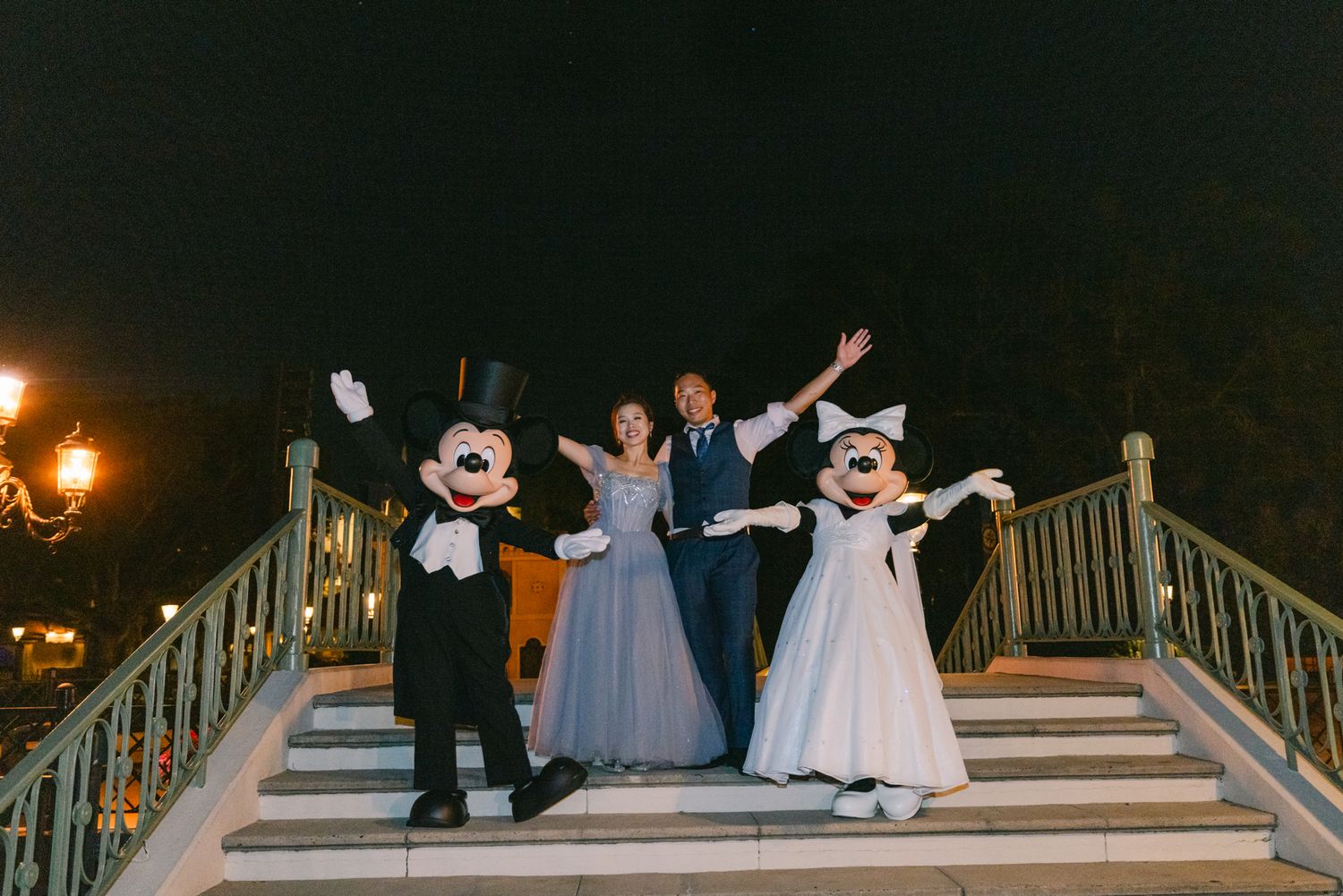 Newlyweds celebrate with Mickey and Minnie Mouse in formal wear on Disney World staircase at night.