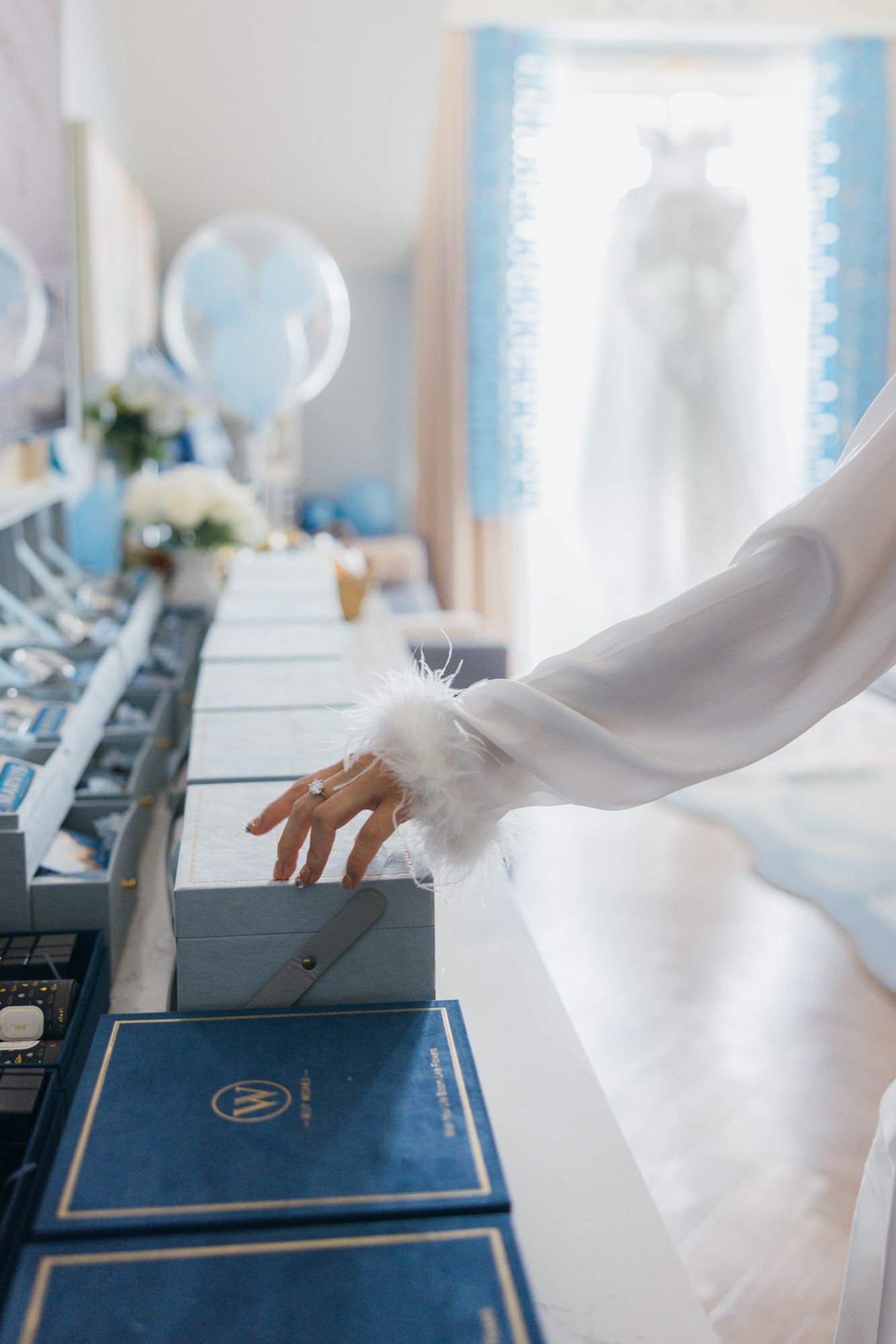 Bride in white feather-trimmed robe opening blue gift boxes arranged on a table at a wedding preparation.