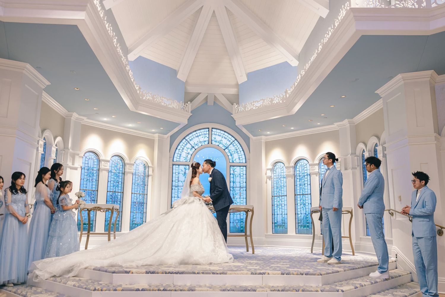 Wedding couple kissing at altar in stunning blue chapel surrounded by bridesmaids and groomsmen.
