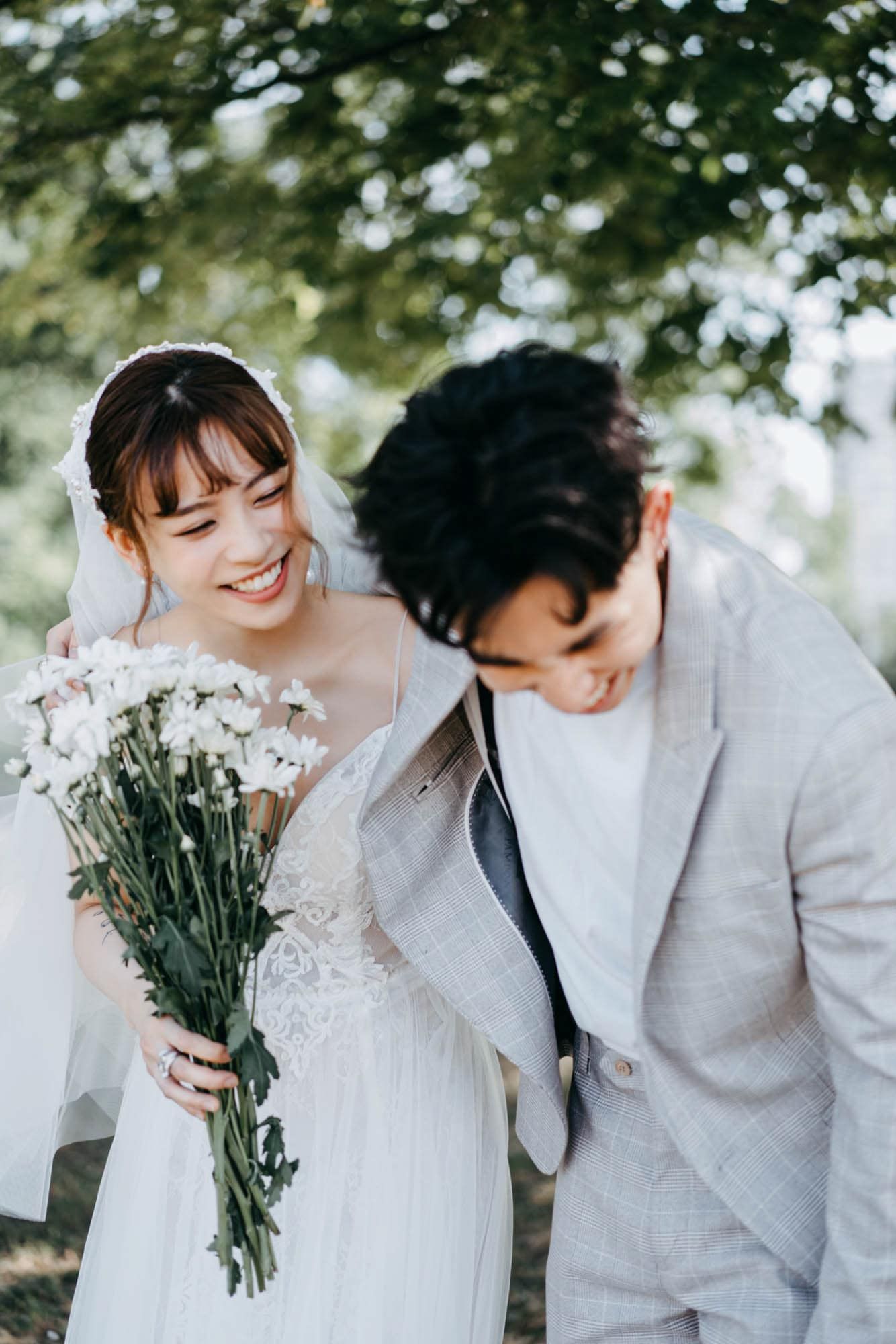 Smiling Asian bride in white lace gown holding white flower bouquet stands beside groom in gray suit outdoors.