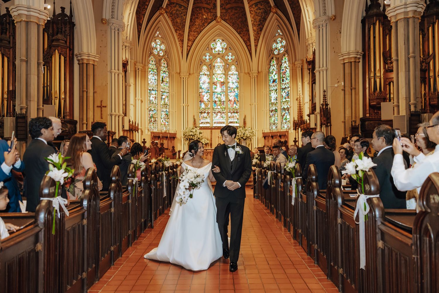 Bride in a white gown and groom in a dark suit walk arm in arm down the church aisle past smiling wedding guests after their ceremony.
