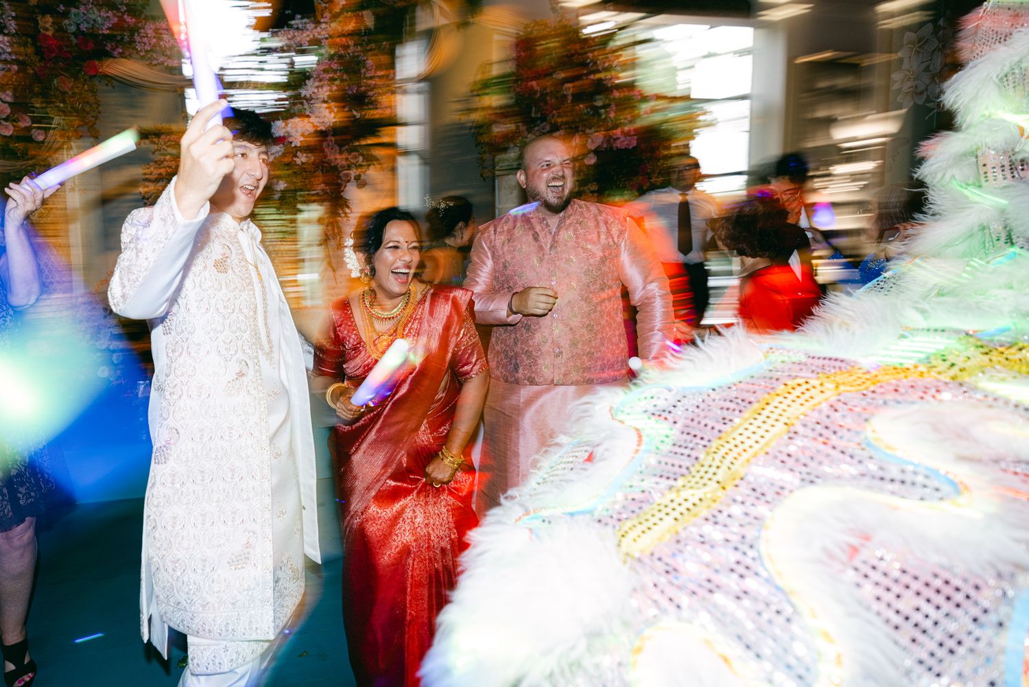 A smiling groom in a cream sherwani and a bride in a red sari celebrate with guests as a white and gold lion dance costume passes by.