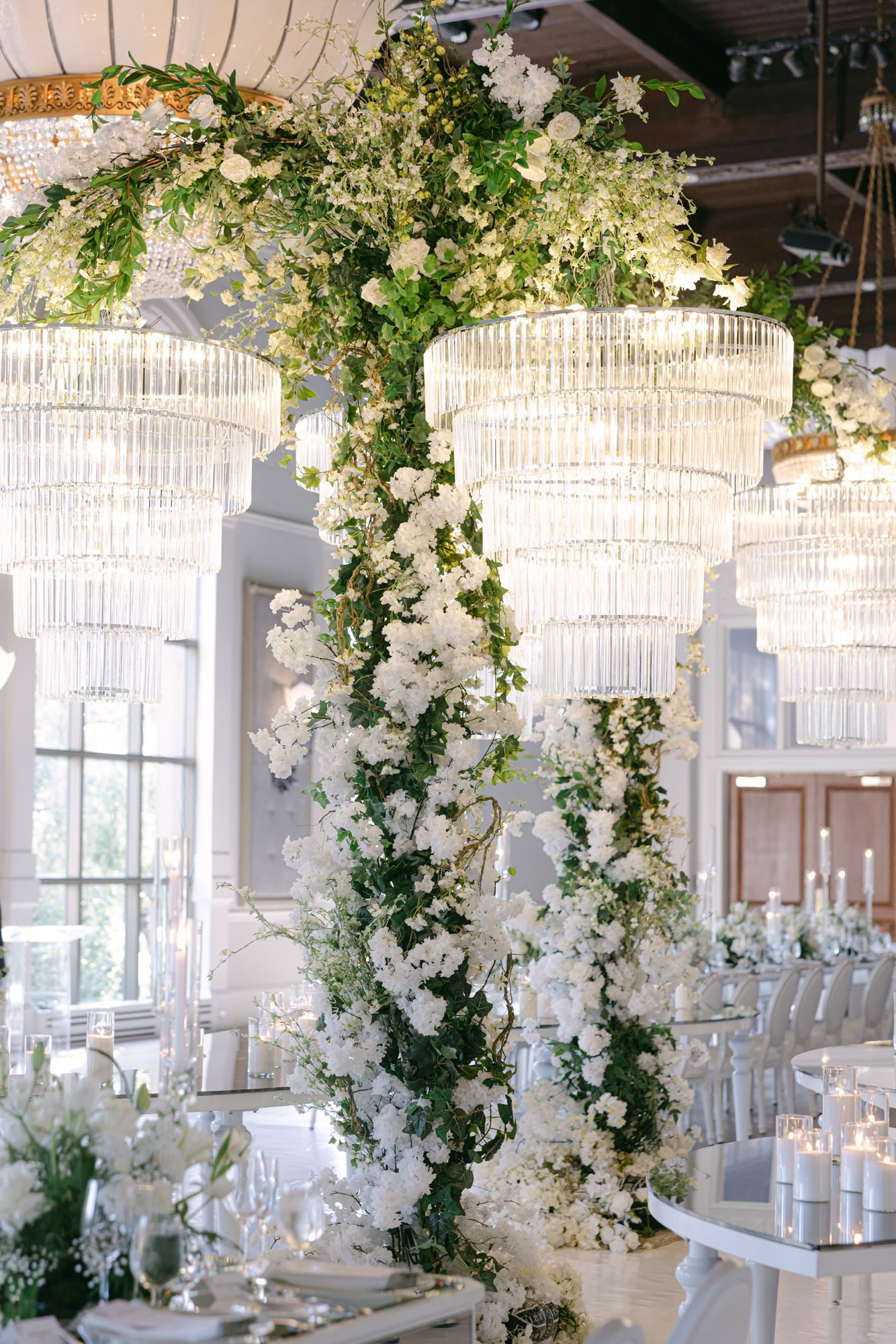 Lush white floral and greenery installation draped around crystal chandeliers above elegant reception tables with candles.