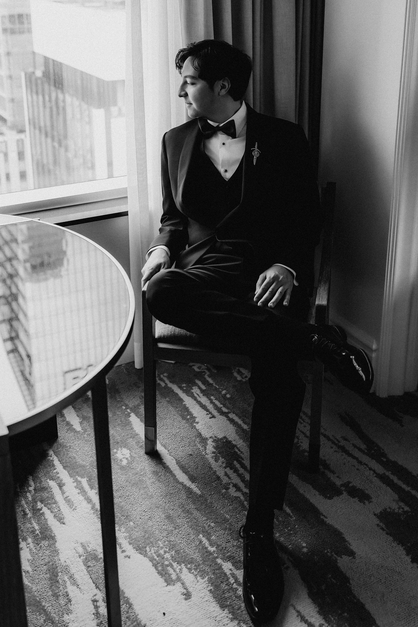 Black and white portrait of a groom in a tuxedo and bow tie seated by a hotel window, gazing outside.