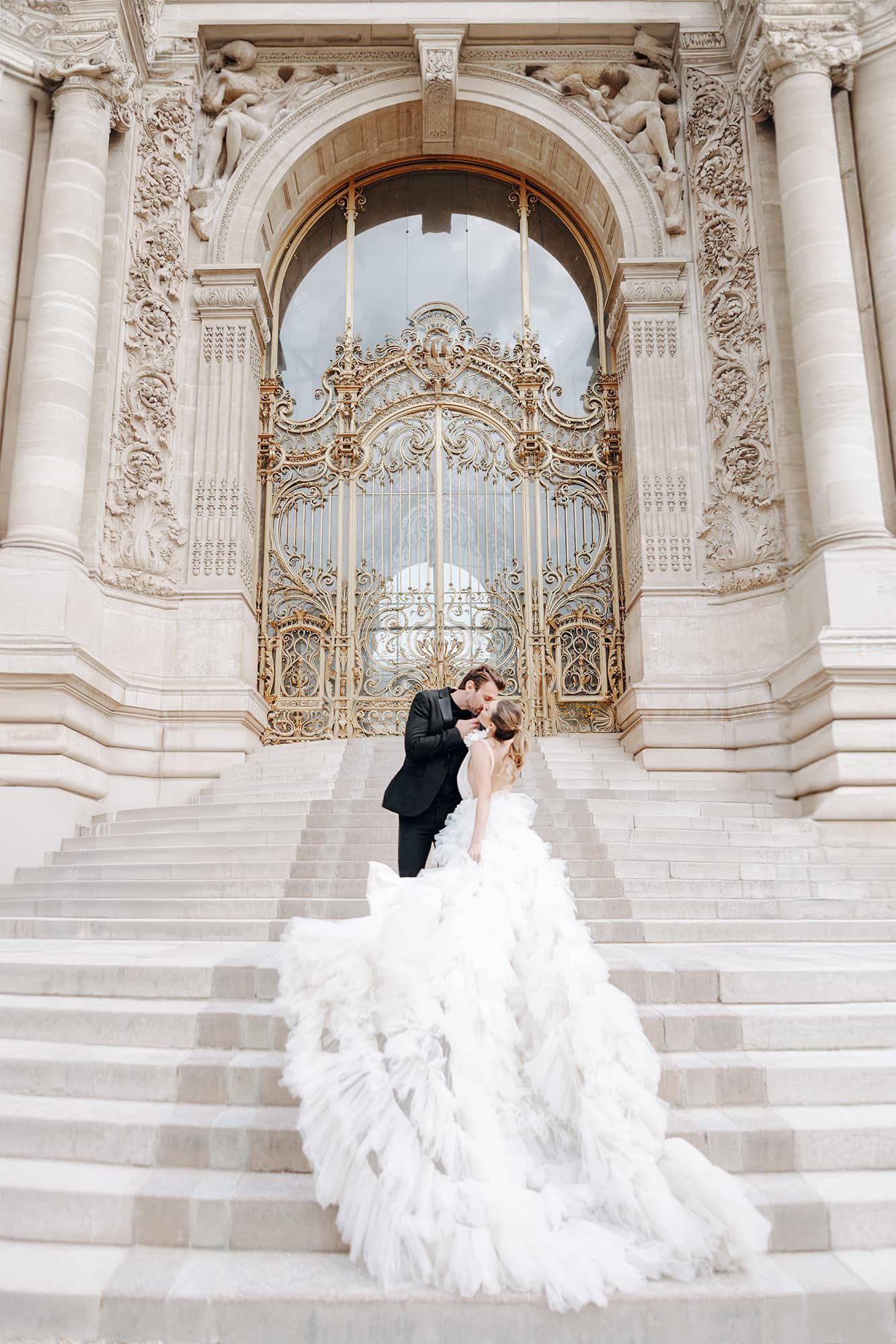 Bride in dramatic ruffled gown and groom kissing on grand steps before ornate golden palace doors.