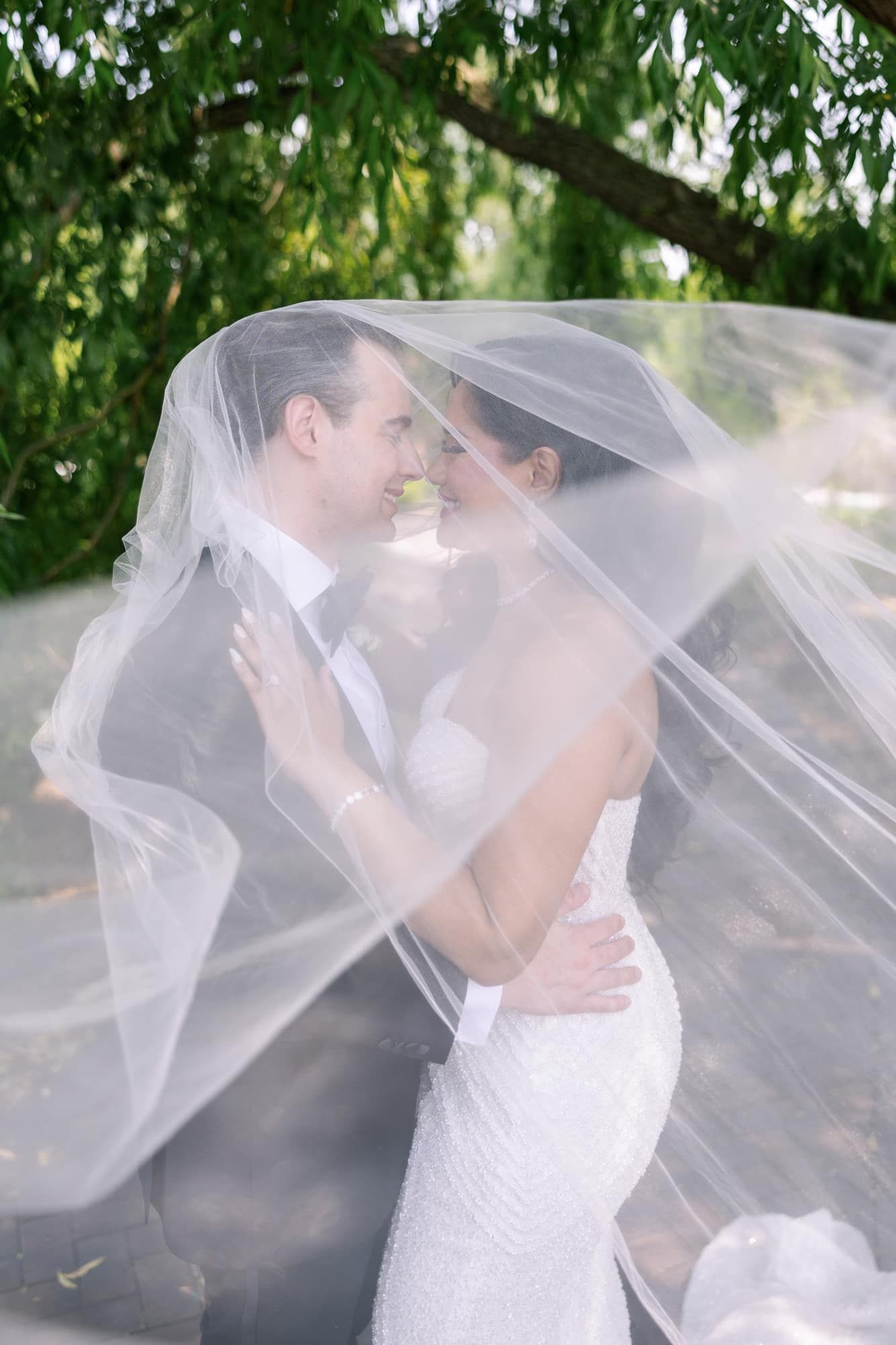 Bride and groom sharing romantic moment under flowing white veil in lush garden setting.