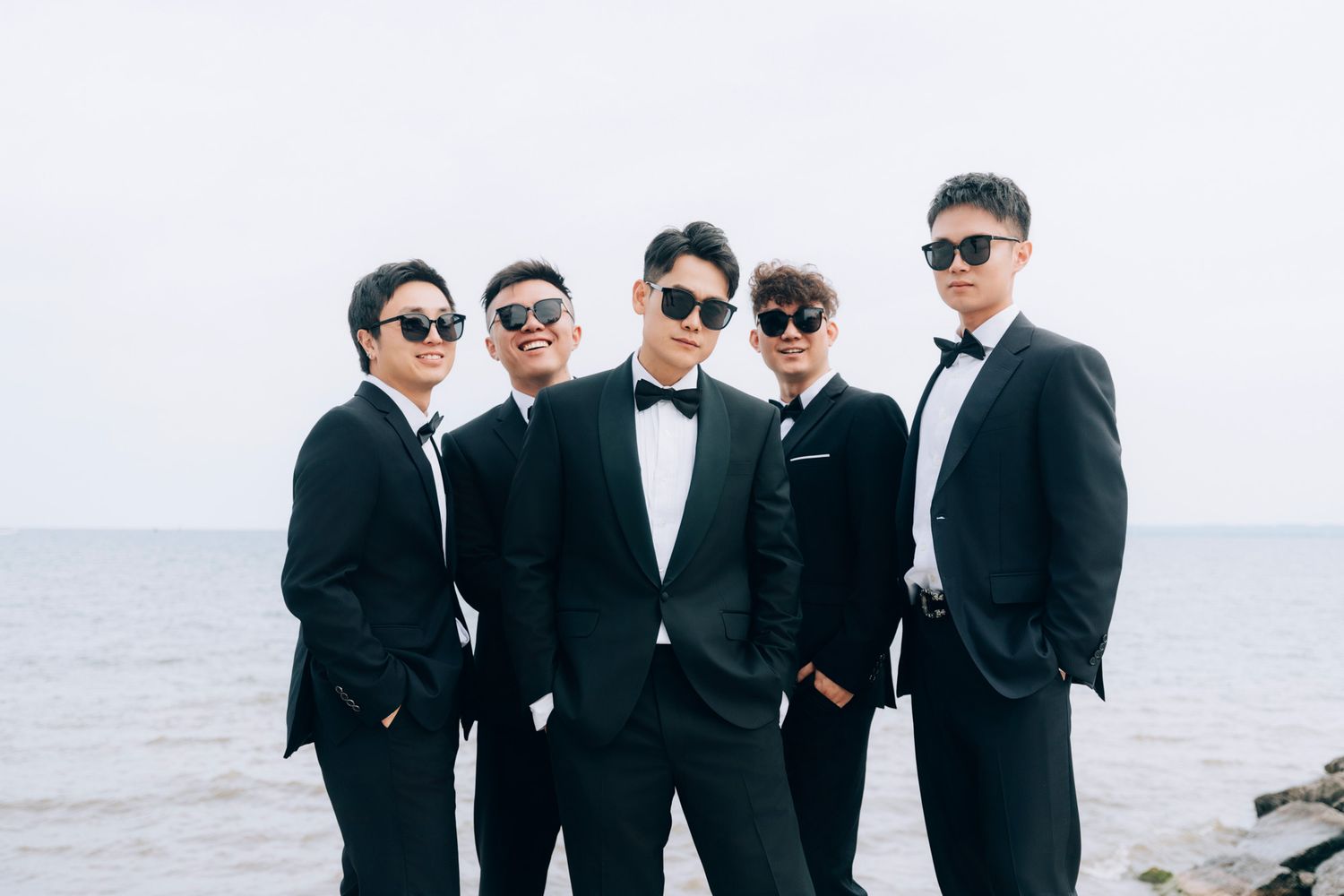 Five groomsmen in black tuxedos and sunglasses pose confidently together by the ocean.