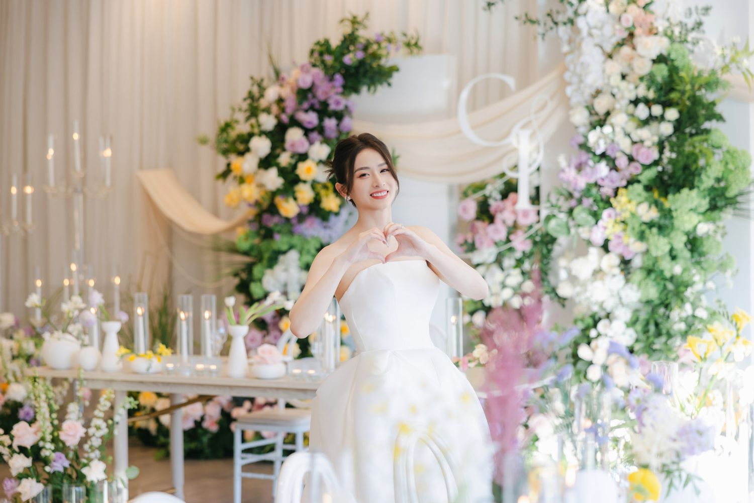 Bride in white strapless gown posing among lush floral wedding decorations with greenery.