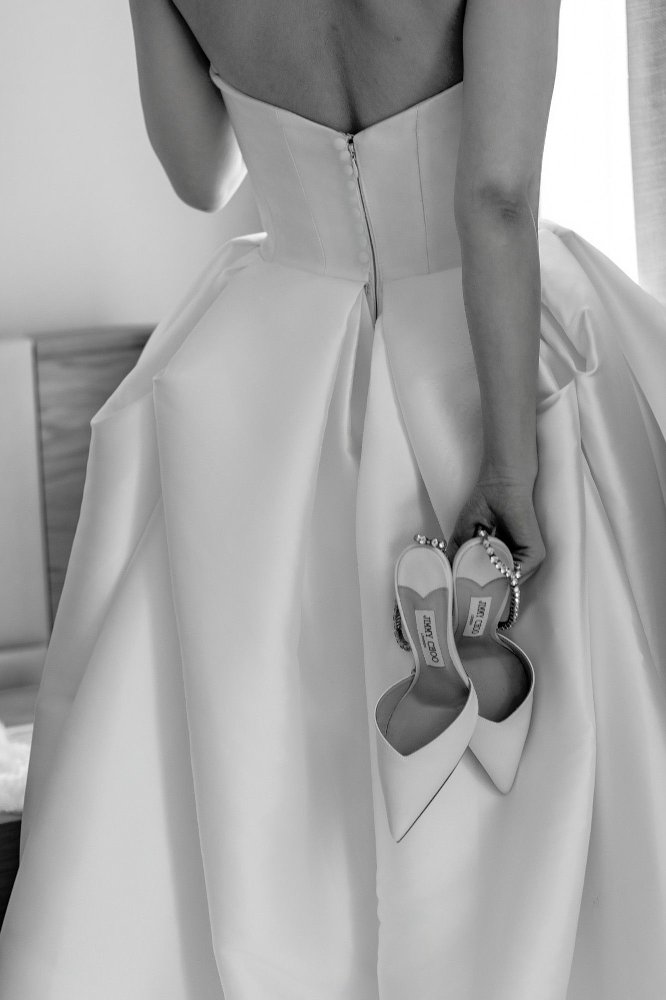 Bride in strapless white wedding gown holding designer heels behind her back, black and white photo.