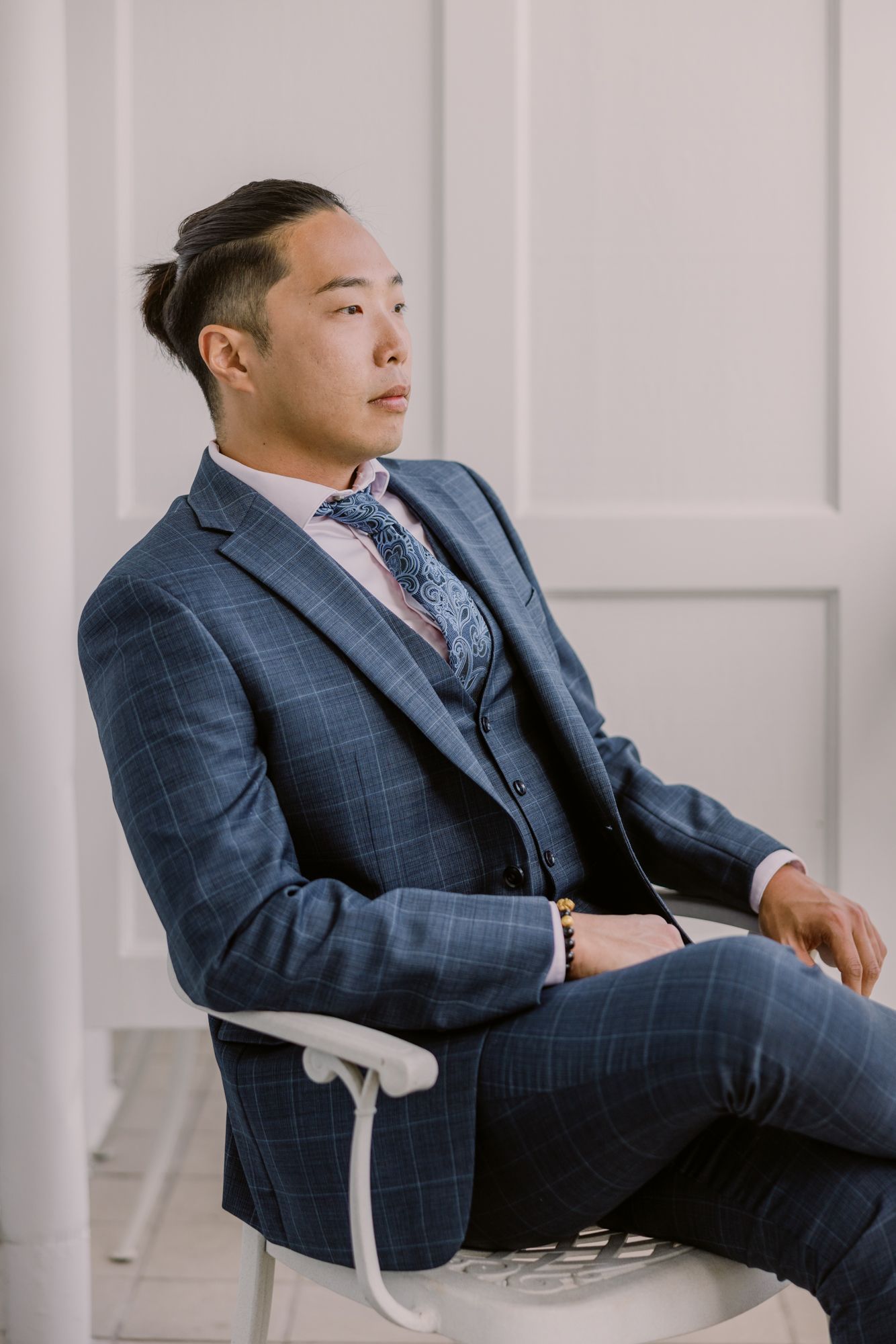 Elegant Asian groom in navy checkered three-piece suit sitting on white chair with blue floral tie.