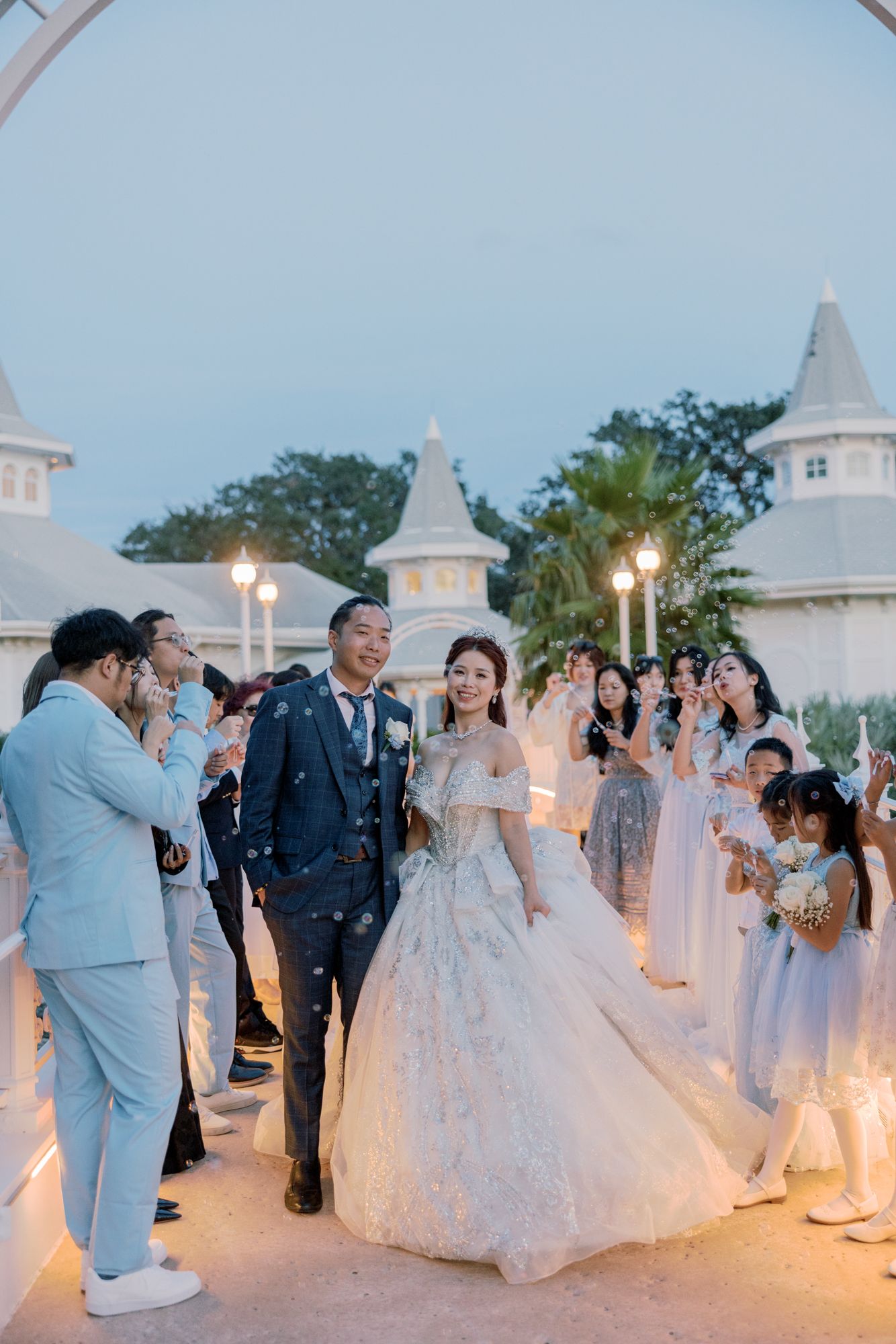 Bride and groom walk through cheering guests after outdoor wedding ceremony at dusk near white chapel.