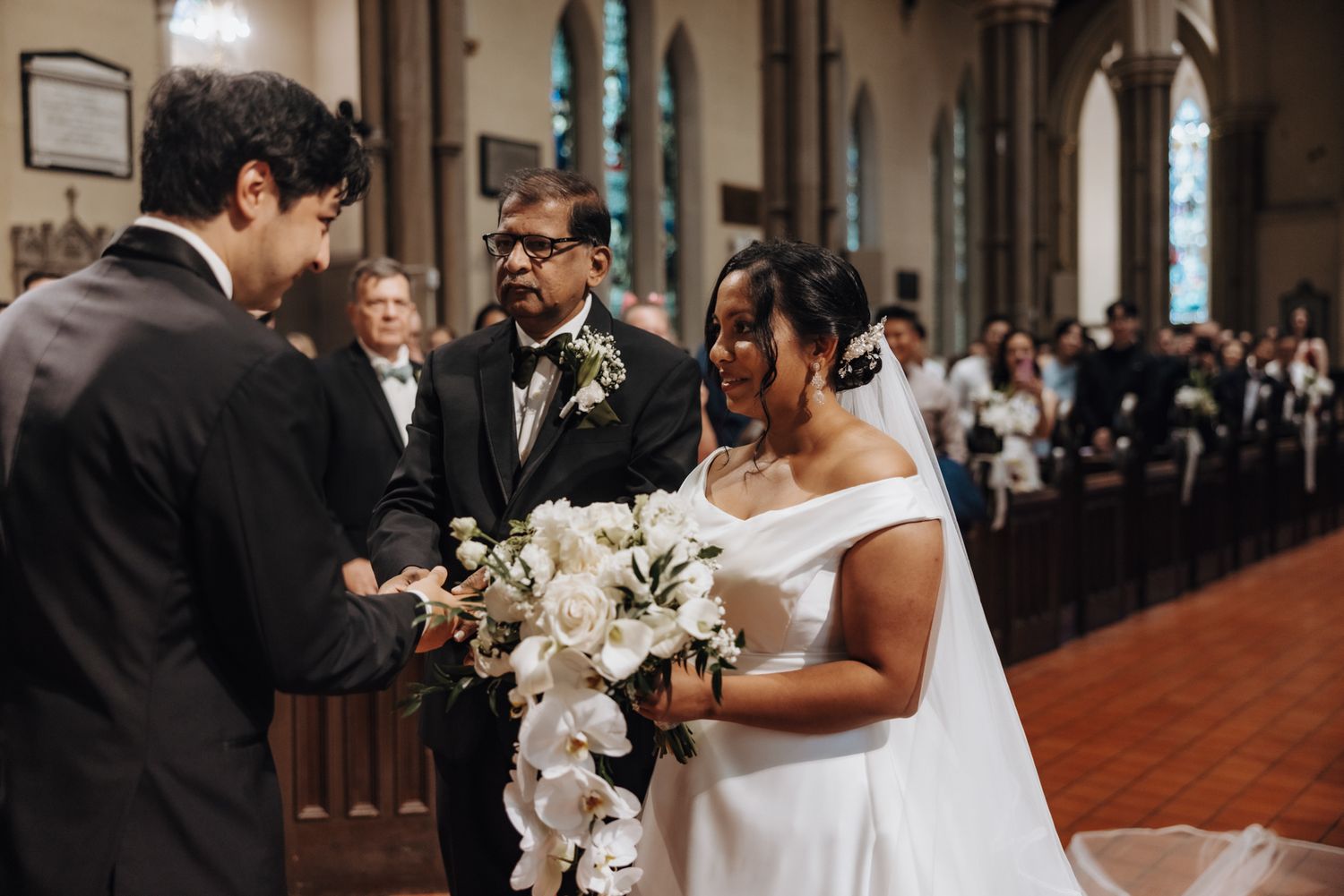 A bride in a white off-shoulder gown holds a white floral bouquet as she joins hands with two men at the church ceremony as guests watch.
