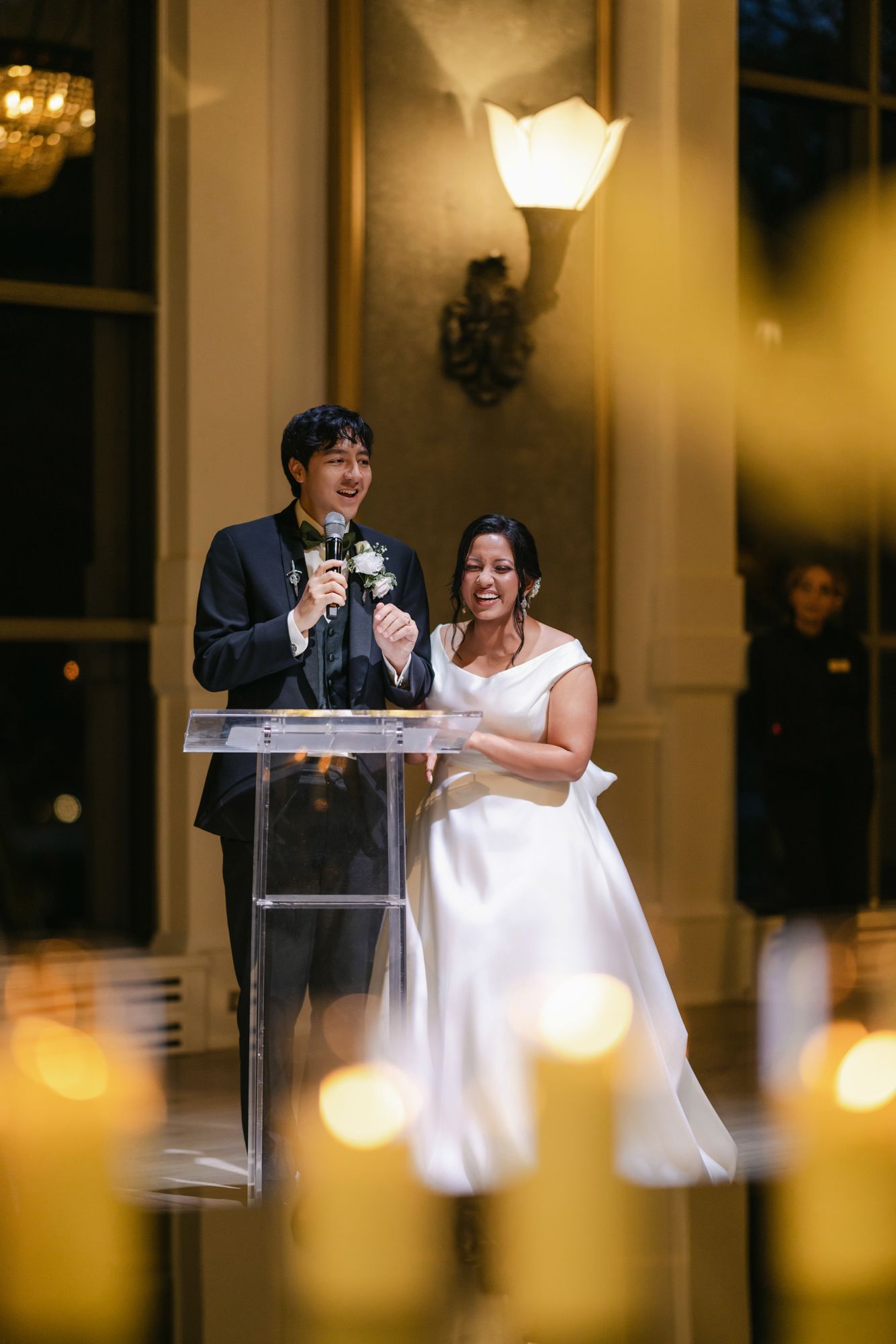 A smiling bride in a white gown and groom in a dark suit give a toast at a glass podium surrounded by glowing candles.