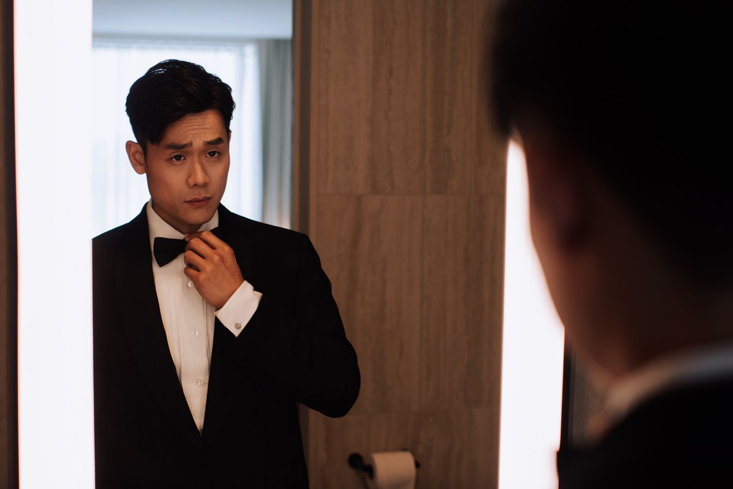 Groom in black tuxedo adjusting bow tie while looking in a mirror.