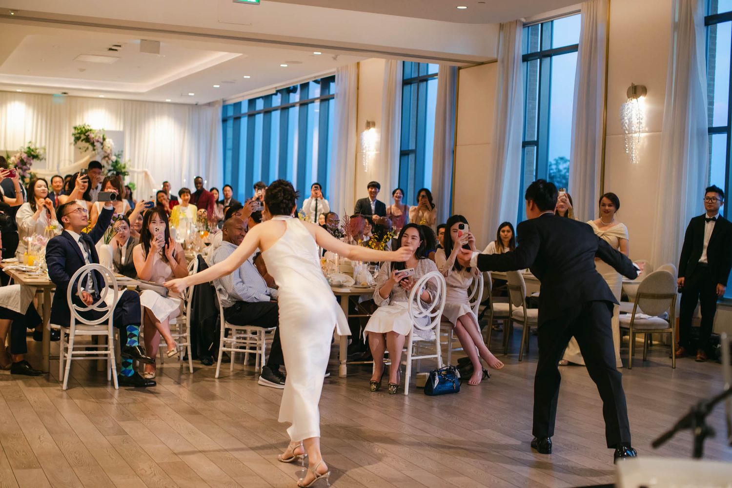 Bride and groom share a lively first dance while wedding guests cheer and applaud in an elegant reception venue.