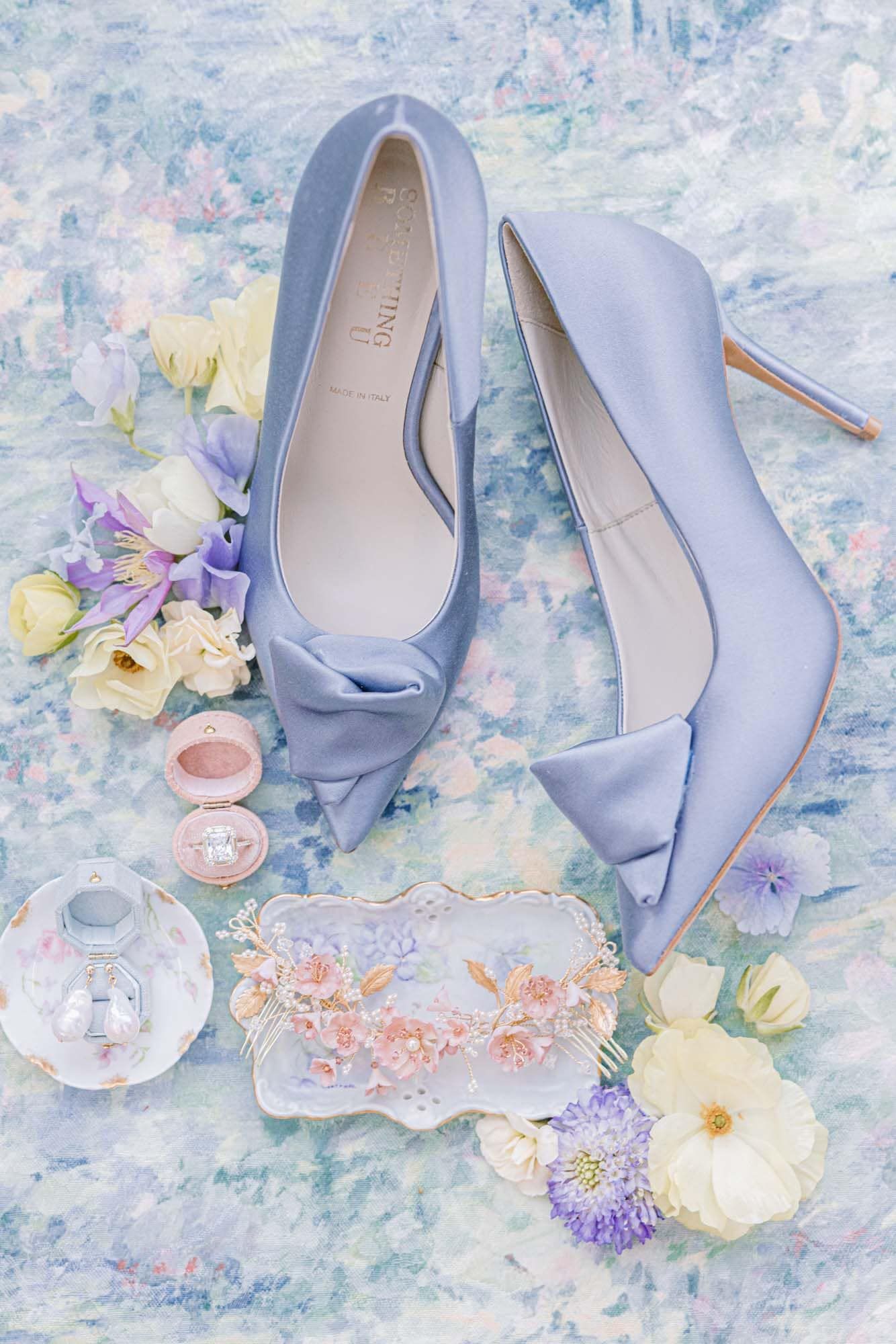Light blue satin bridal heels with bow detail surrounded by flowers, perfume, and ring box on floral background.