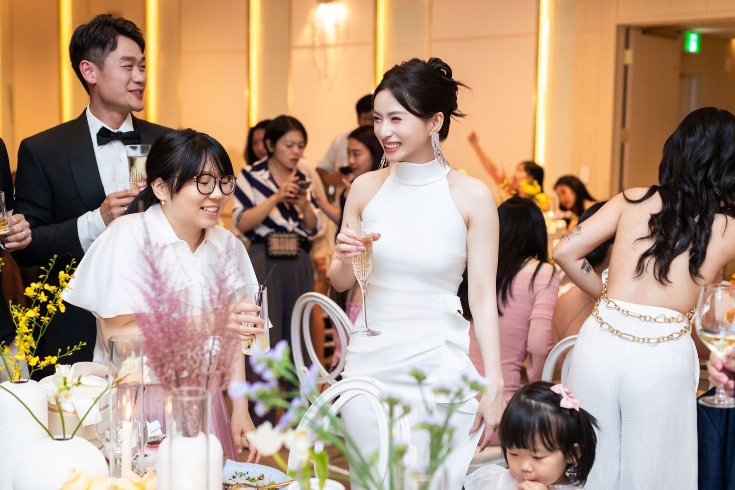 Radiant bride in white halter gown smiling with young guests at elegant wedding reception.