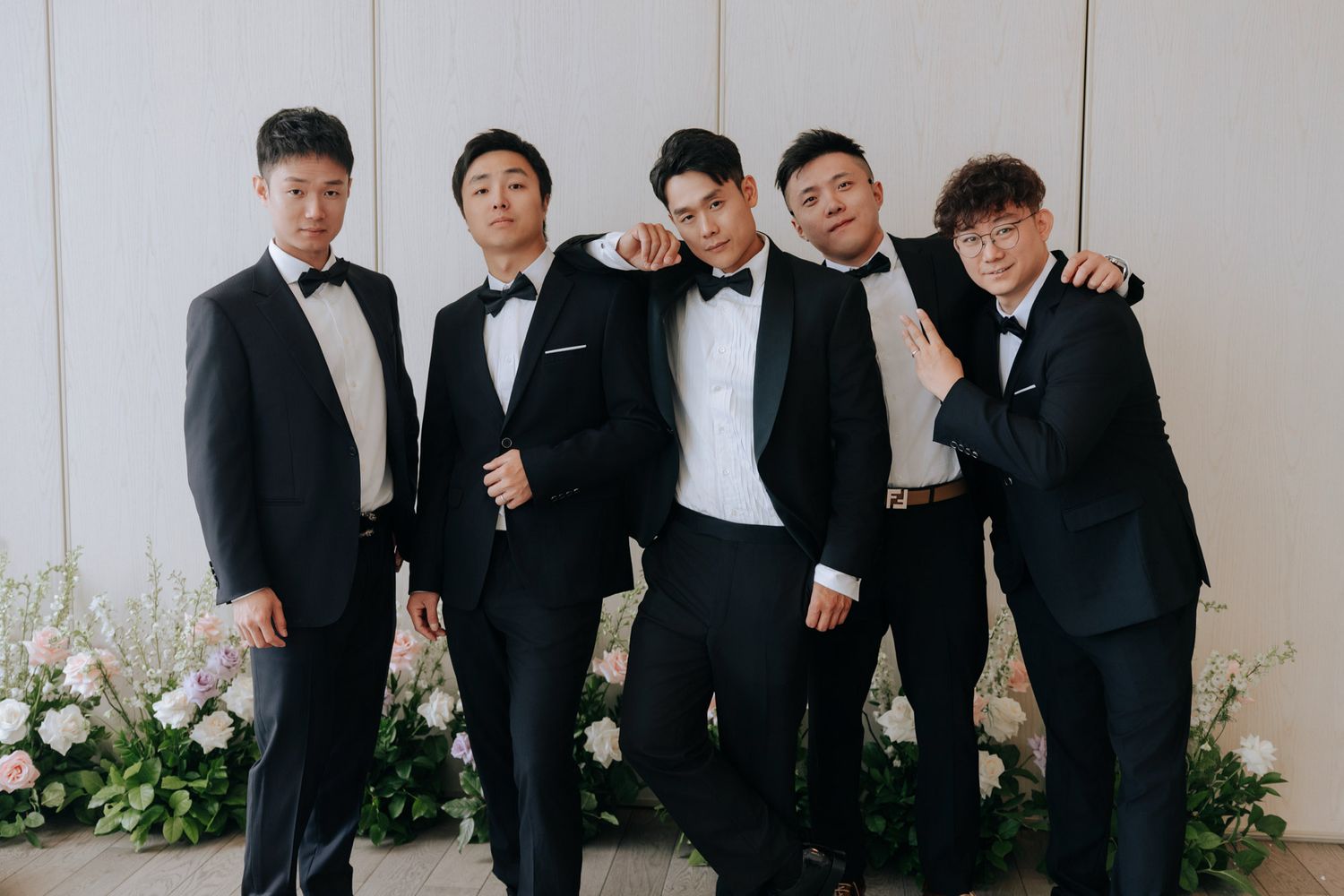 Five groomsmen in black tuxedos pose together smiling at a wedding reception with floral arrangements.