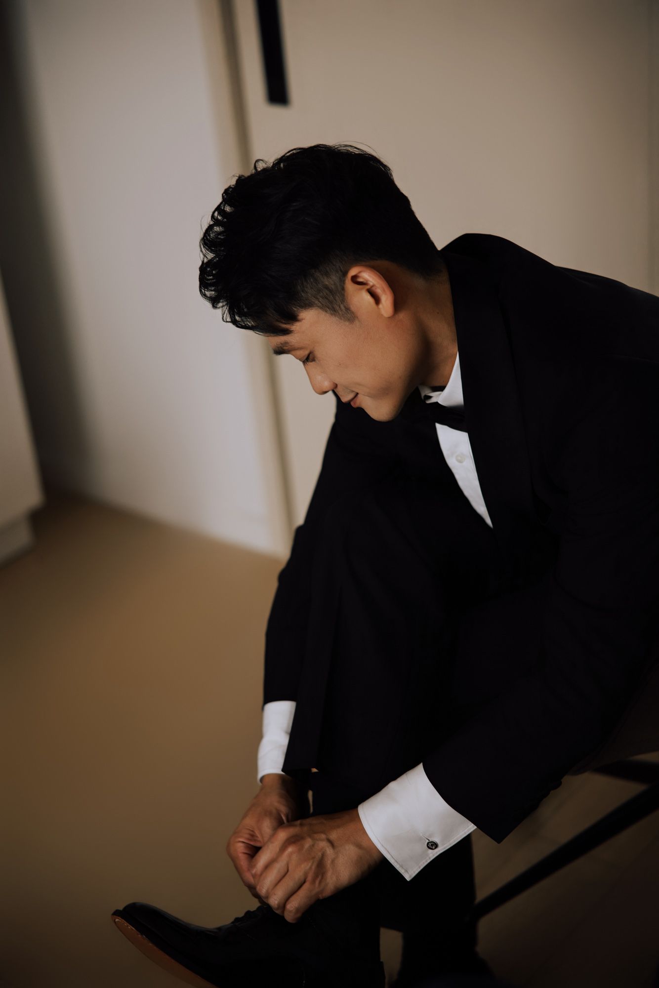 Dapper groom in black tuxedo and bow tie putting on dress shoes before wedding.