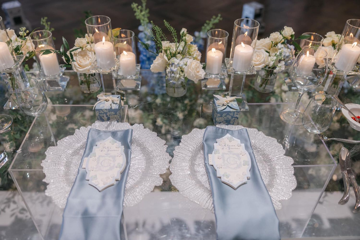 Elegant wedding place setting with white doily chargers, blue satin napkins, and candles on a mirrored table.