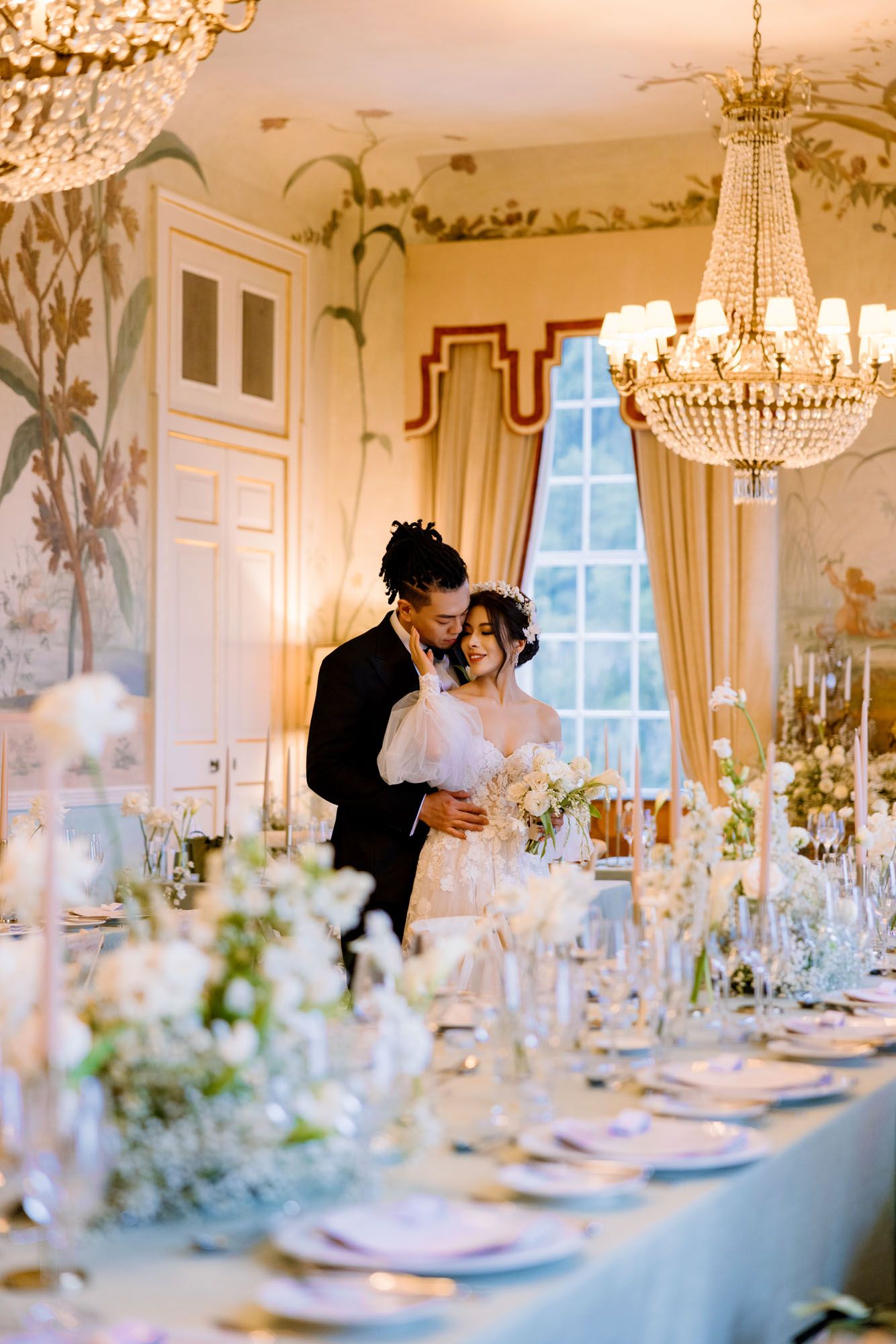 Bride and groom embrace in elegant chandelier-lit reception hall with white floral table settings.