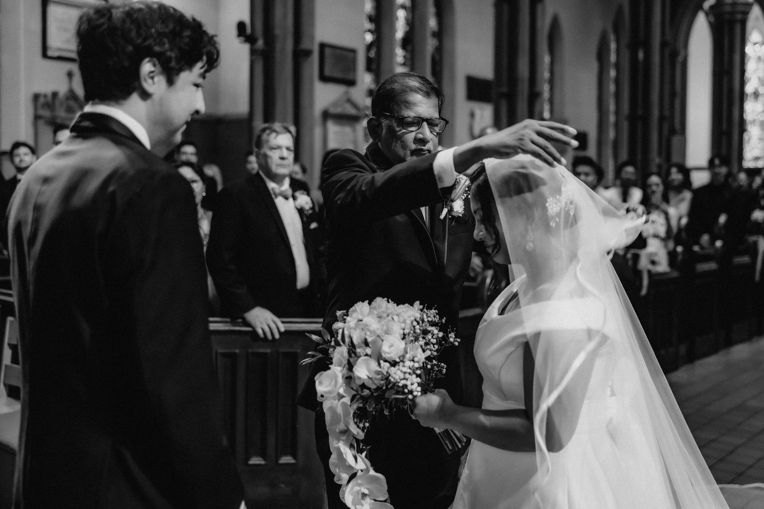 Black and white photo of a father lifting the bride's veil at the church altar as the groom looks on.