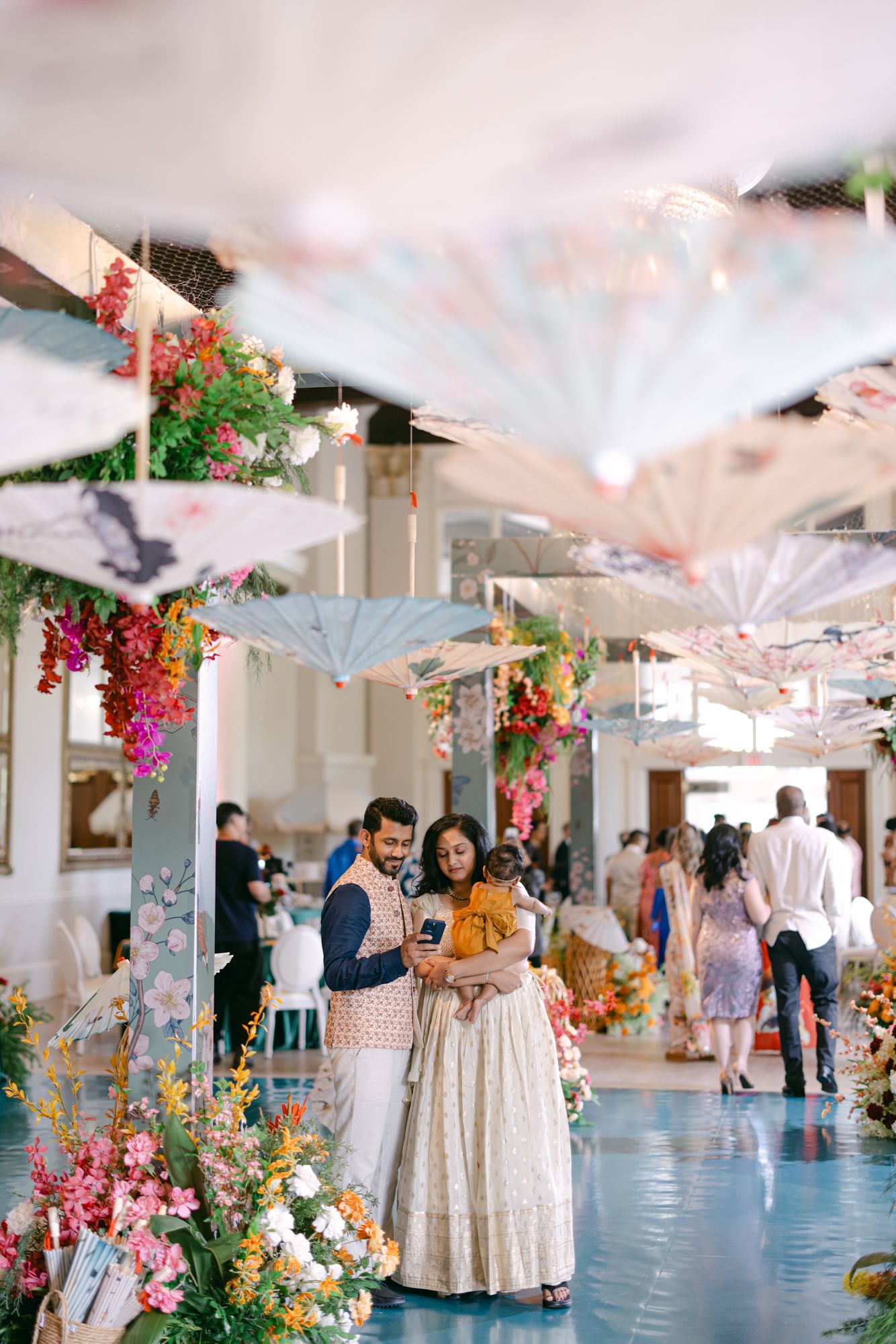 A couple in traditional Indian wedding attire look at a phone beneath hanging decorative umbrellas and bright floral arrangements.