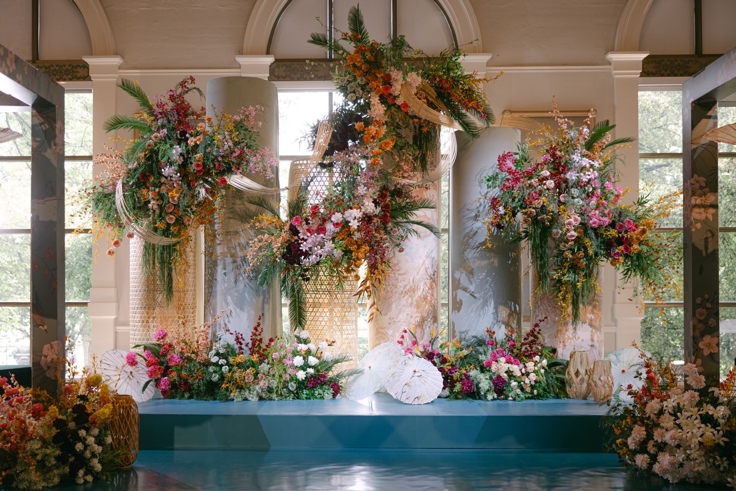 A lavish wedding stage features towering arrangements of pink, orange and green flowers framed by stone columns and tall windows.