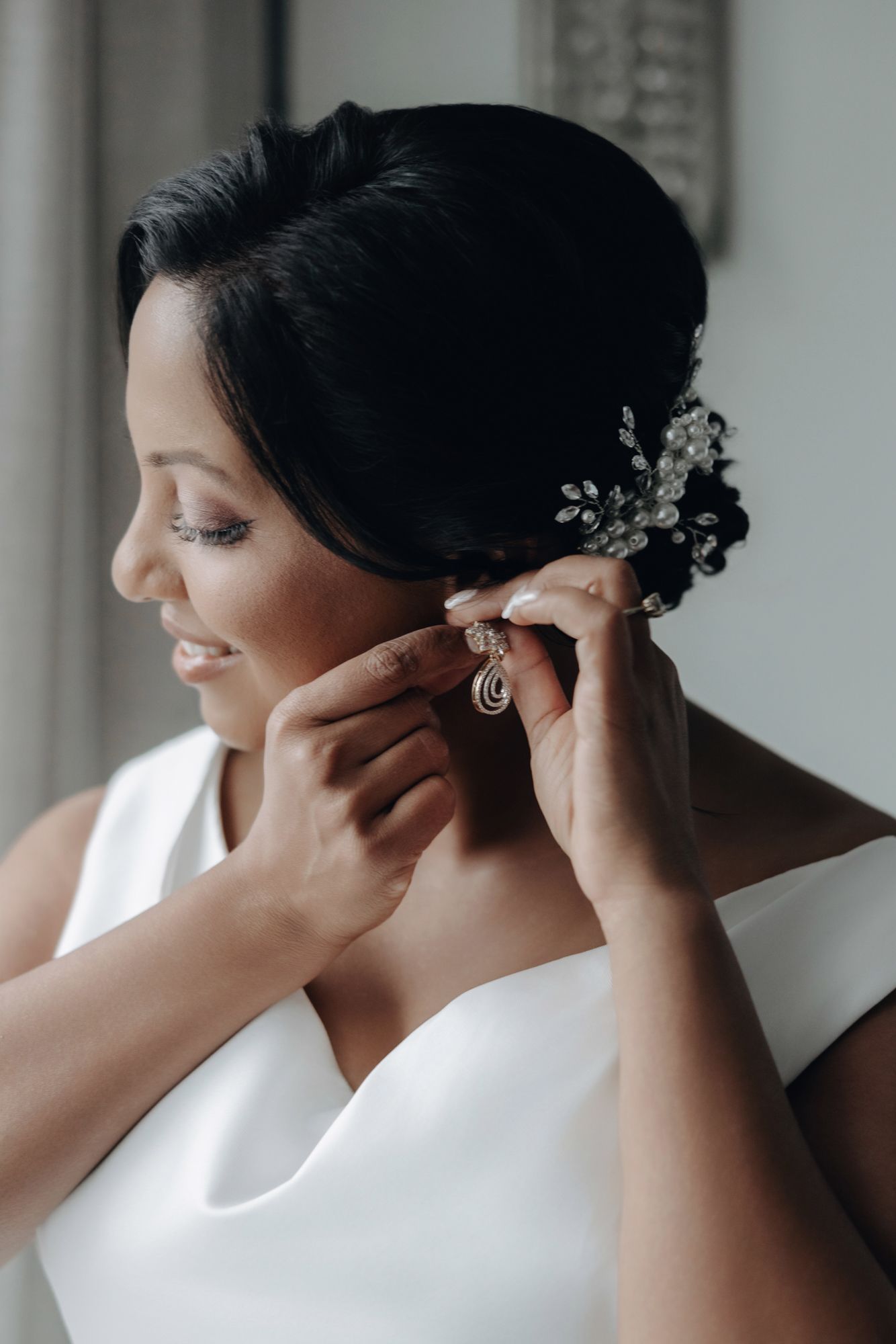 Smiling bride in a white gown with a crystal hair clip puts on a drop earring while getting ready for her wedding.