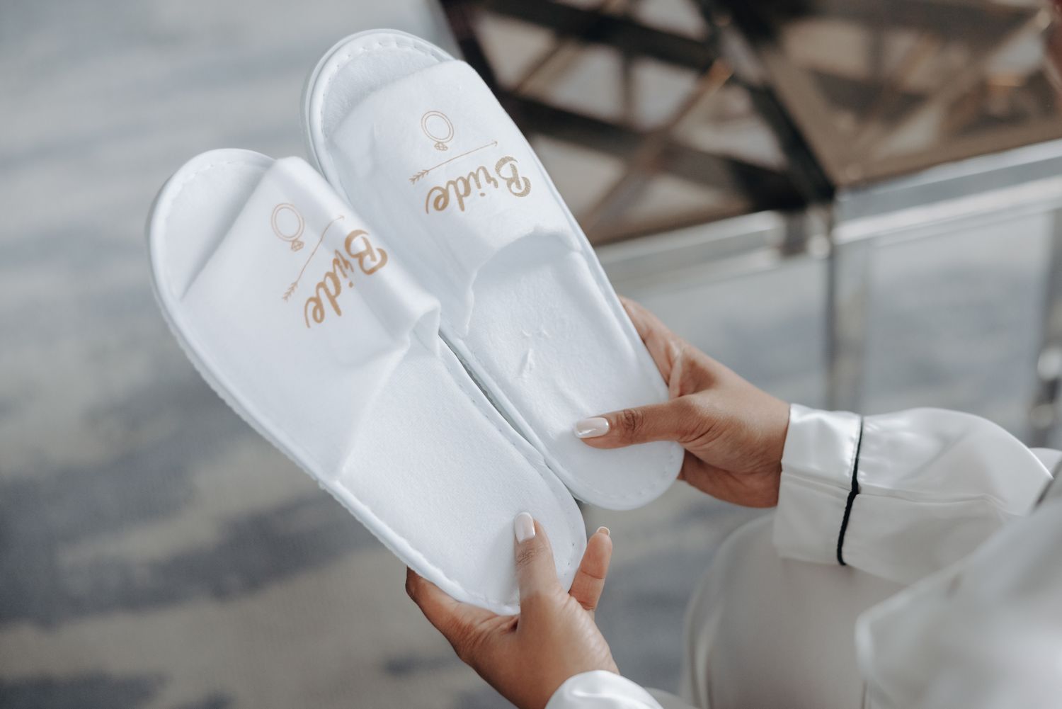 Woman in a white satin robe holds a pair of white open-toe bridal slippers embroidered with the word Bride in gold lettering.