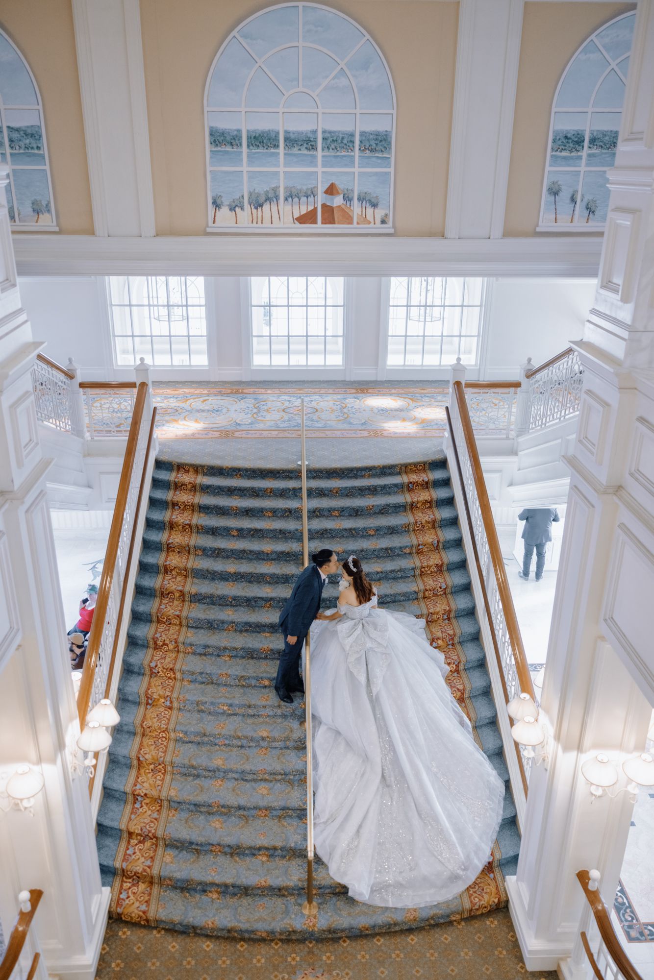 Bride and groom share a kiss on grand staircase in luxurious white wedding venue with high ceilings.