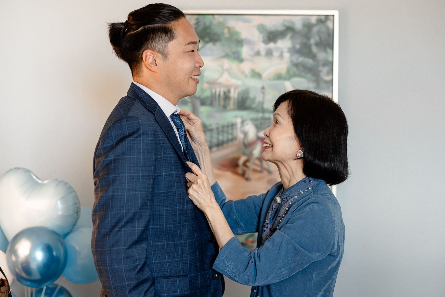 A groom in a blue suit gets his tie adjusted by a smiling woman, with blue heart balloons in background.