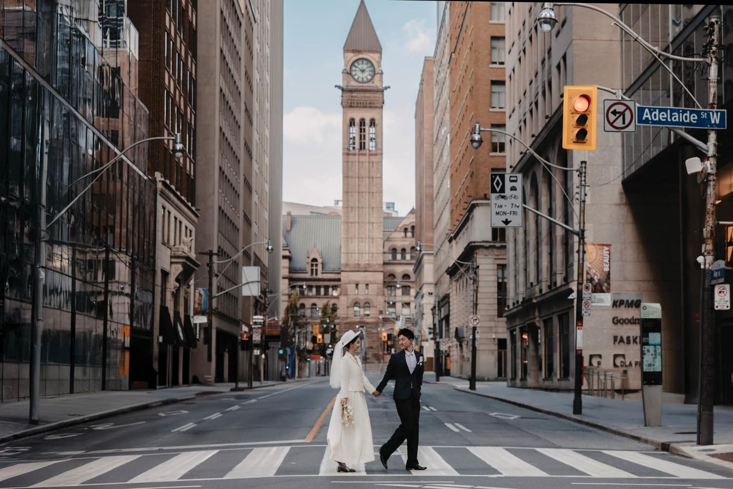Bride and groom cross a zebra crossing on a quiet urban street with a historic clock tower in the background.
