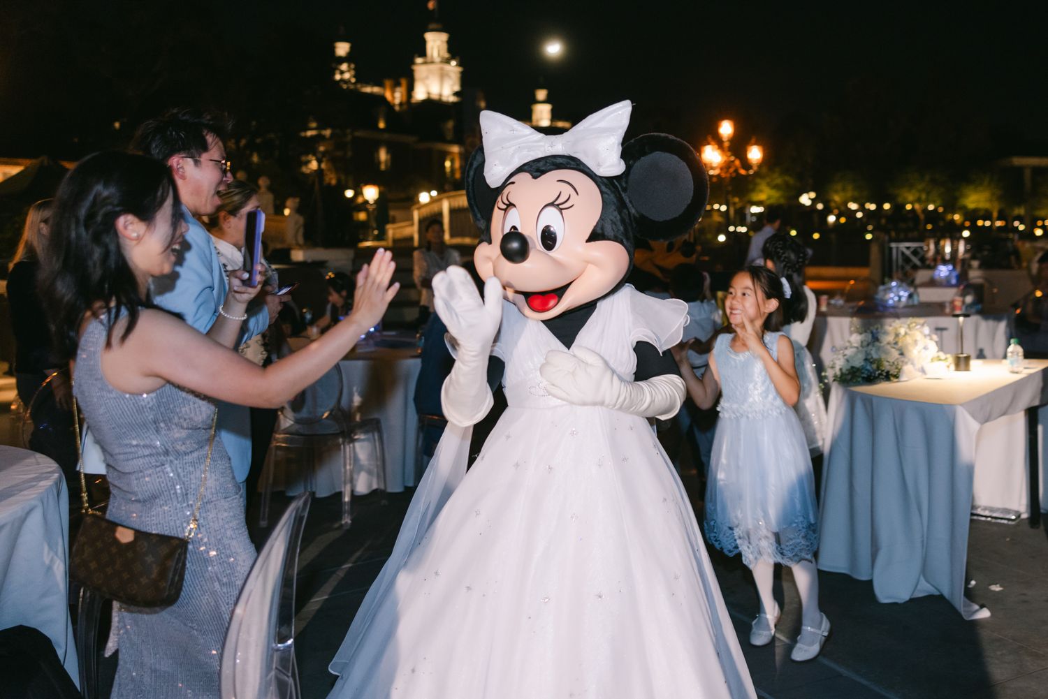 Minnie Mouse in white wedding gown dances with guests including young children at Disney wedding reception.