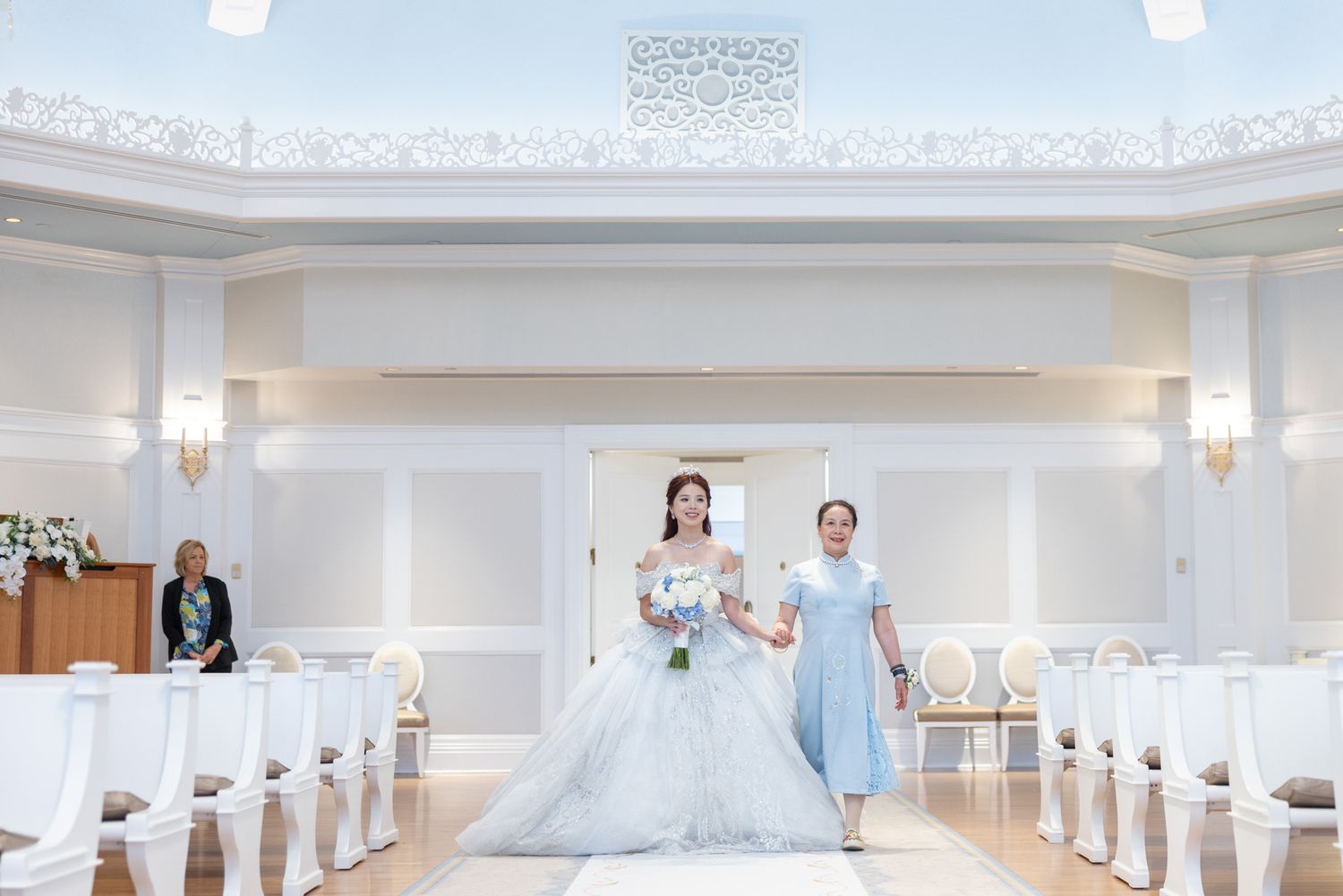 Bride in blue lace gown walking down white chapel aisle accompanied by woman in blue dress.